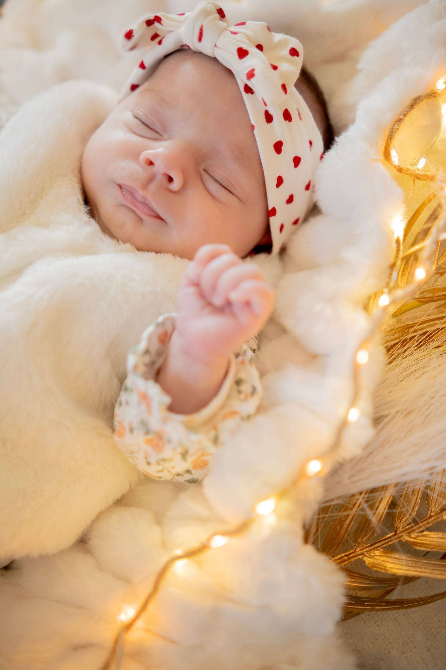 A sleeping newborn nestled in soft white cotton with string lights and a heart-patterned hat creating a dreamy atmosphere.