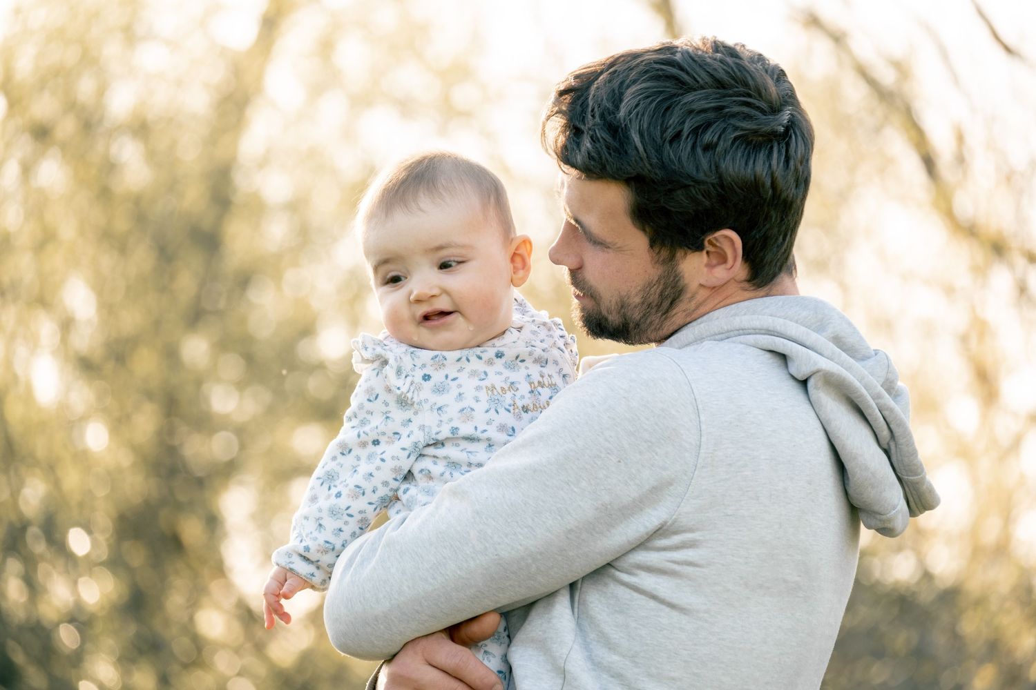 A bearded person in a gray hoodie holds a smiling baby wearing a patterned outfit during a sunny outdoor moment.