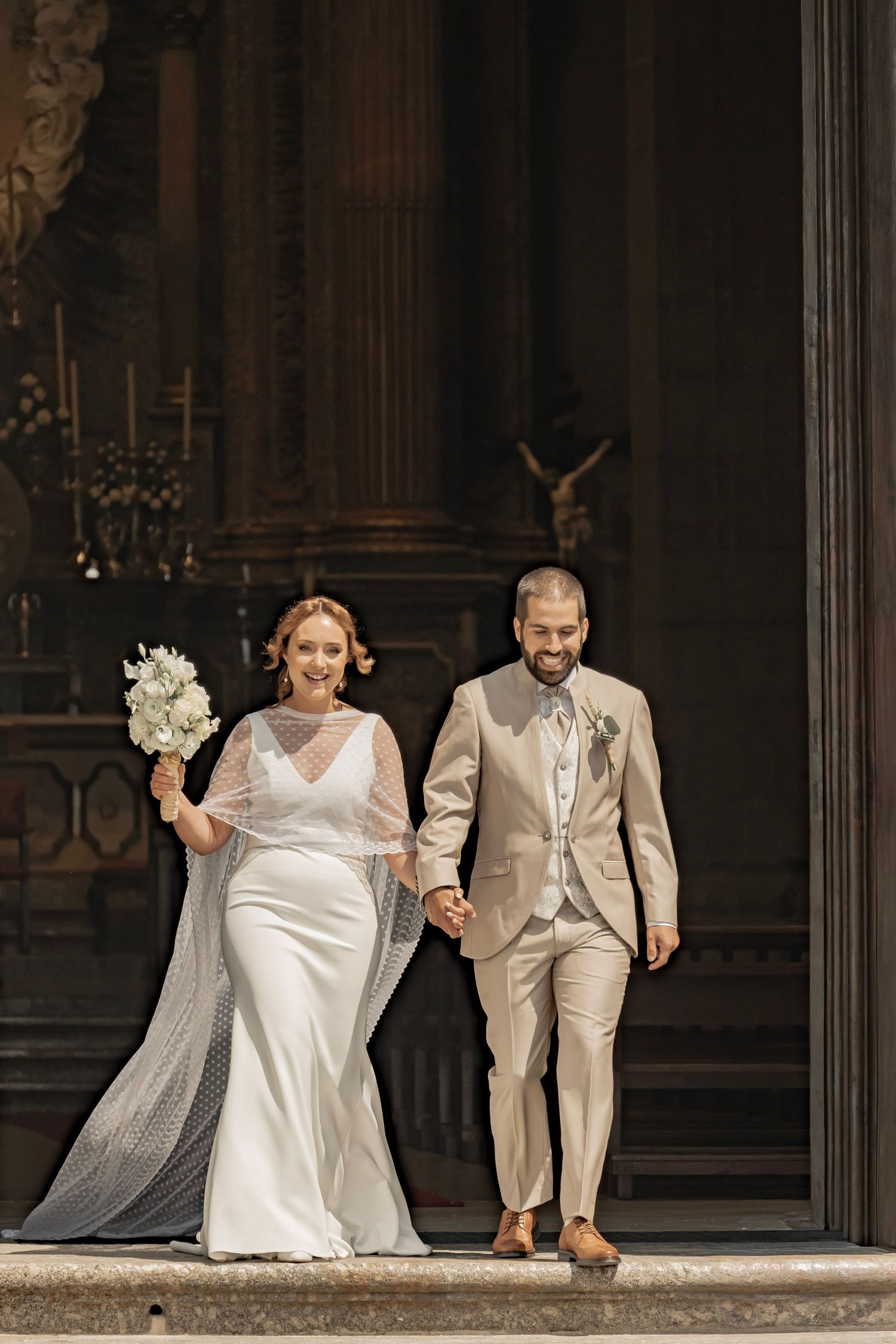 Bride in white gown holding bouquet and groom in beige suit walk hand in hand from a church after wedding ceremony.