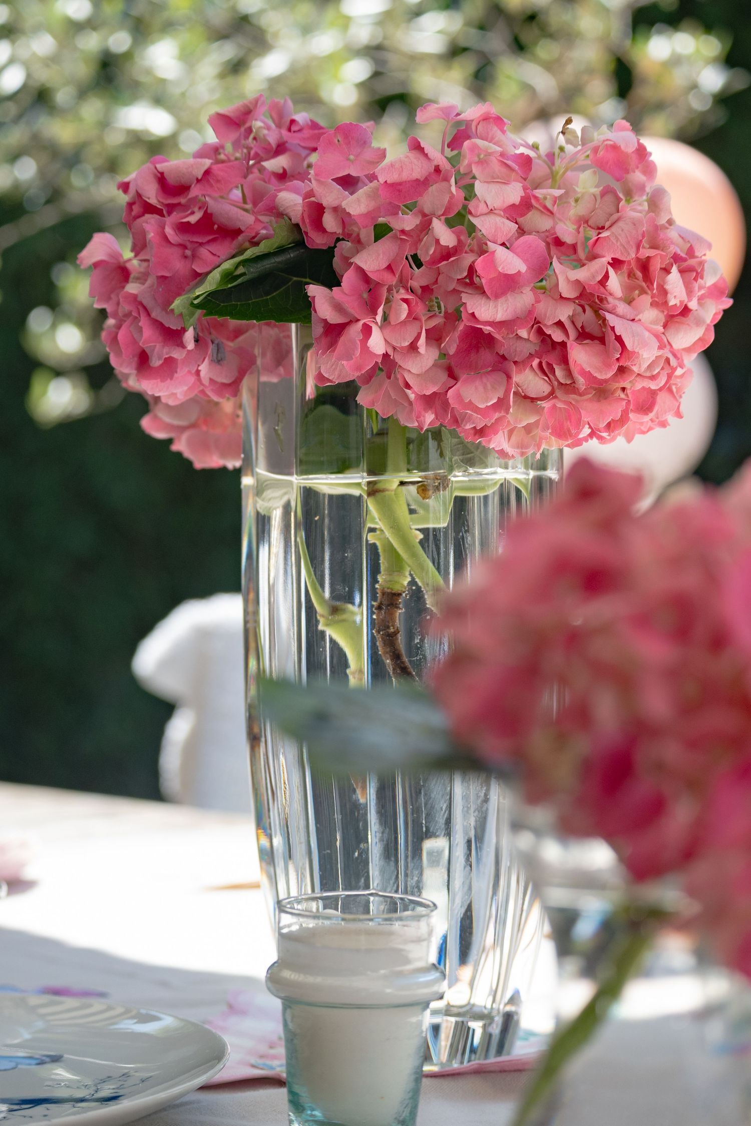 Pink hydrangea bouquet in clear glass vase on outdoor table setting with soft bokeh garden background.