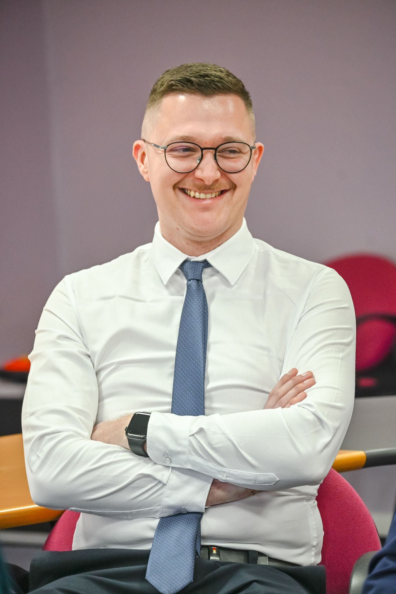 A professional wearing glasses and blue tie smiles confidently with arms crossed.