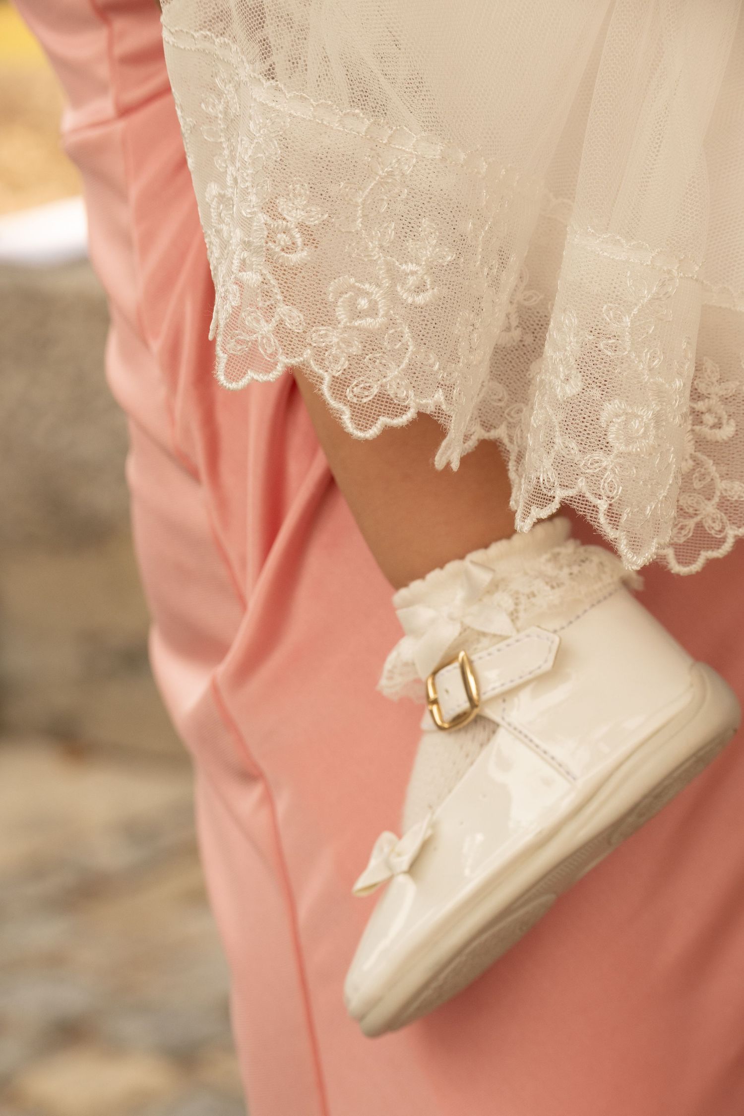 A delicate white lace dress flows gracefully over a soft pink background, with a white shoe peeking out from beneath.