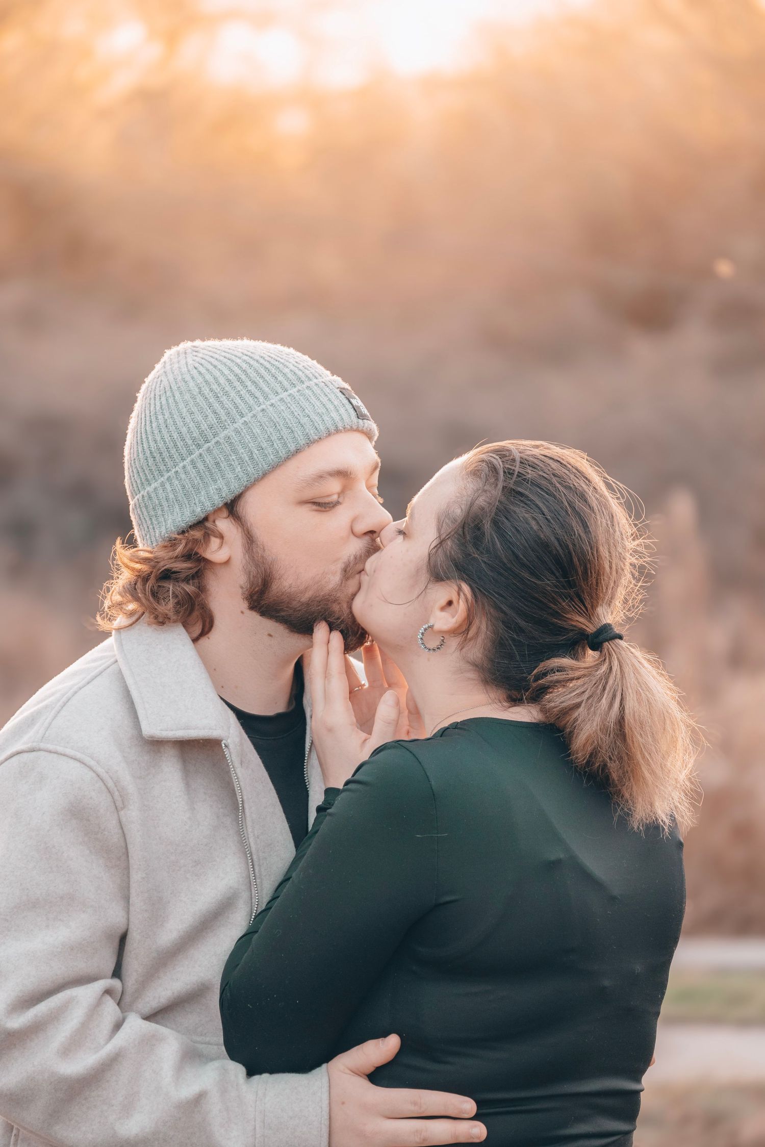 A romantic couple sharing a tender kiss outdoors at sunset with one wearing a winter beanie hat.