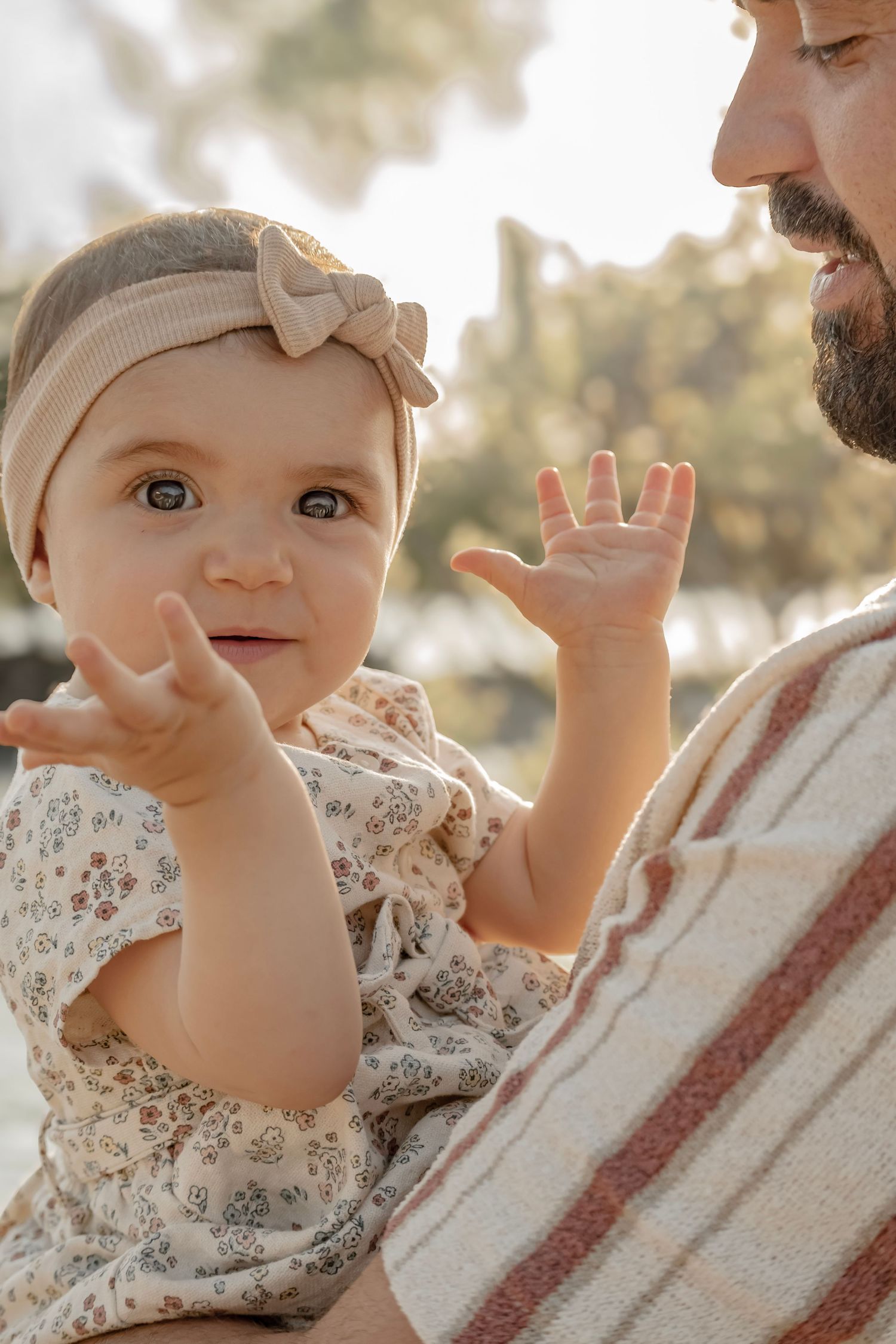 Adorable baby girl in floral outfit and bow headband being held by bearded father outdoors in warm sunlight.