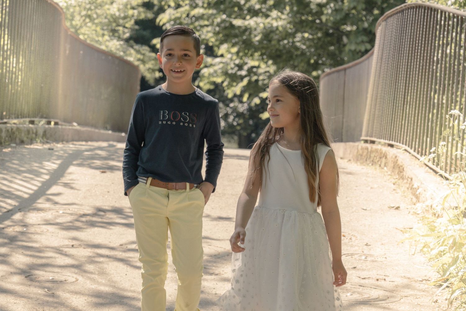 Two young people walking together on a path, one wearing a navy sweater and khakis, the other in a white dress.