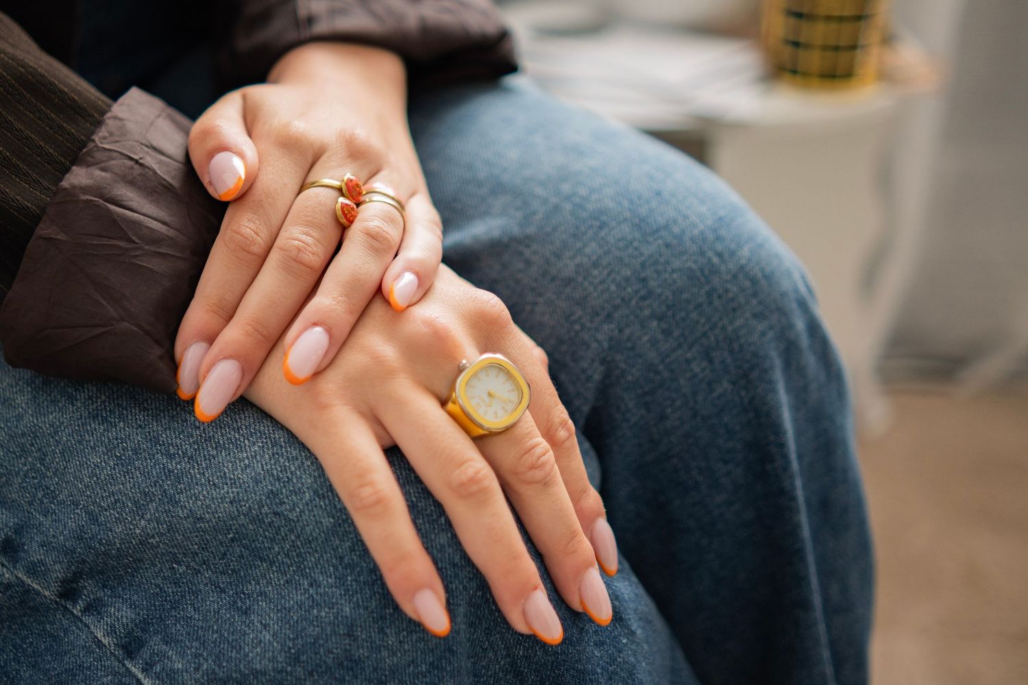 Close-up of hands with gold rings and a watch, featuring nude pink nails, resting on blue denim jeans.