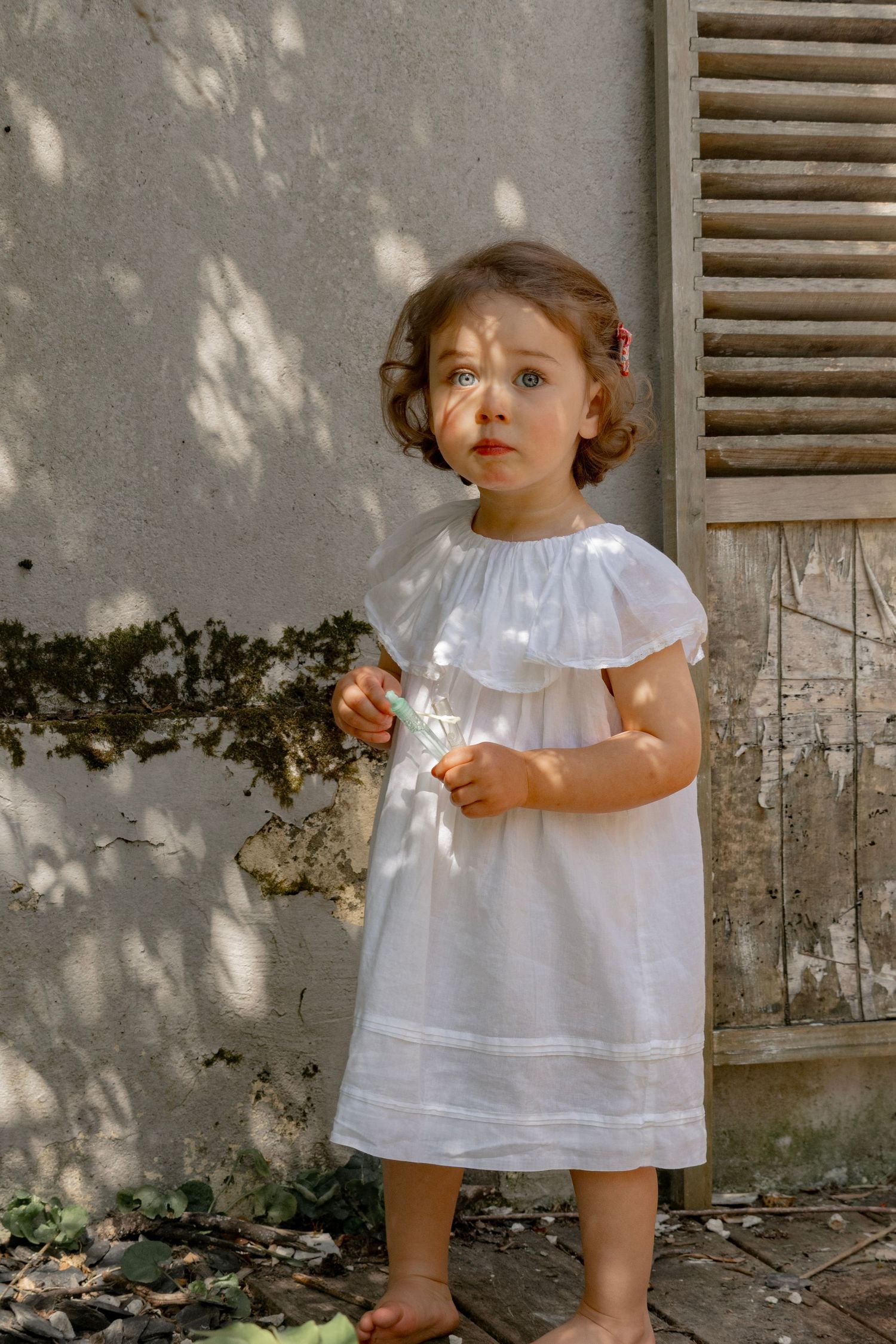 Toddler girl in white ruffle dress standing barefoot outdoors near a rustic wall with dappled sunlight.