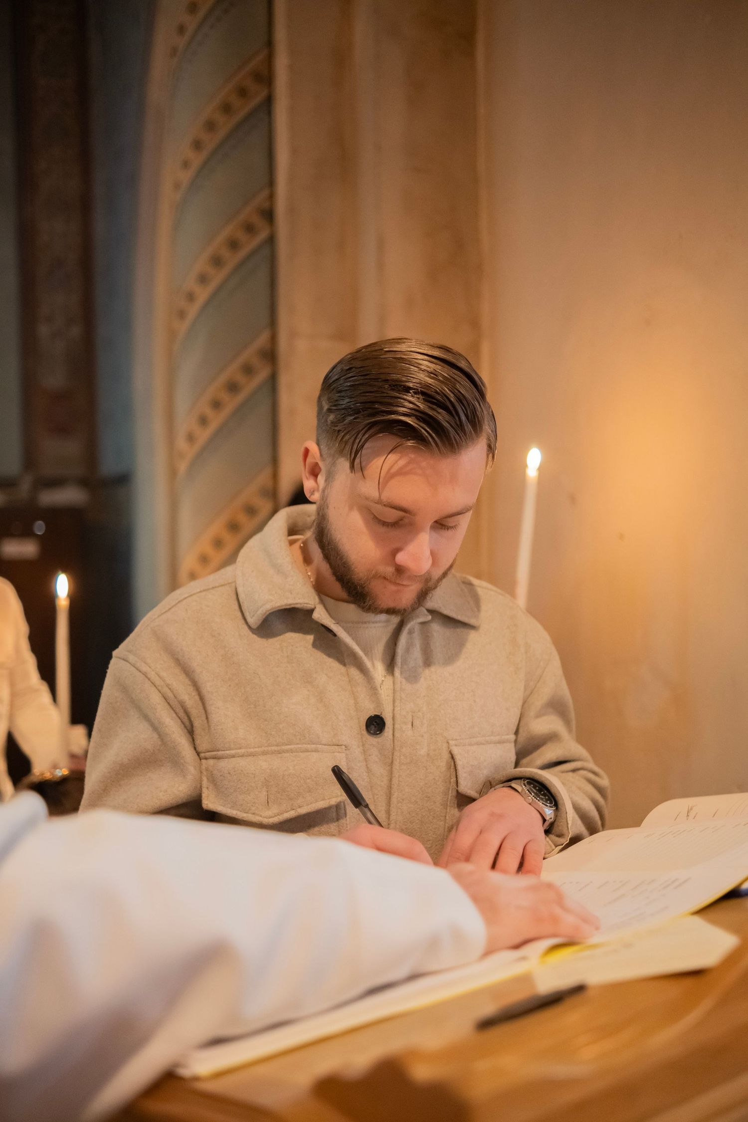 A person writes in a book with a fountain pen in a warm, candlelit setting with elegant architectural details.