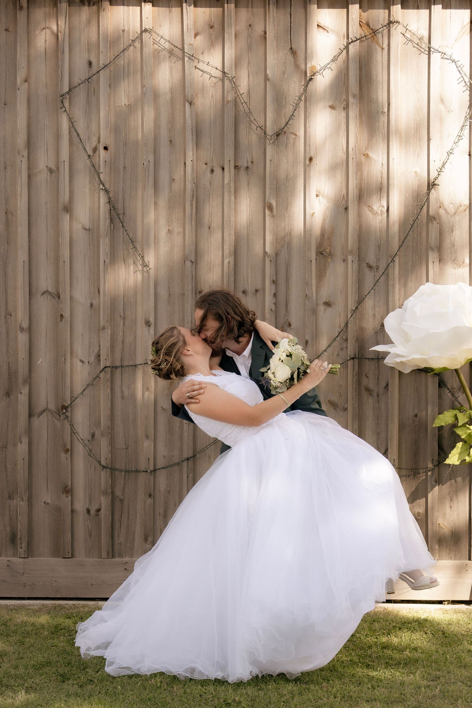 Bride and groom share a romantic dip kiss in front of a rustic wooden barn wall, bride holding white bouquet.