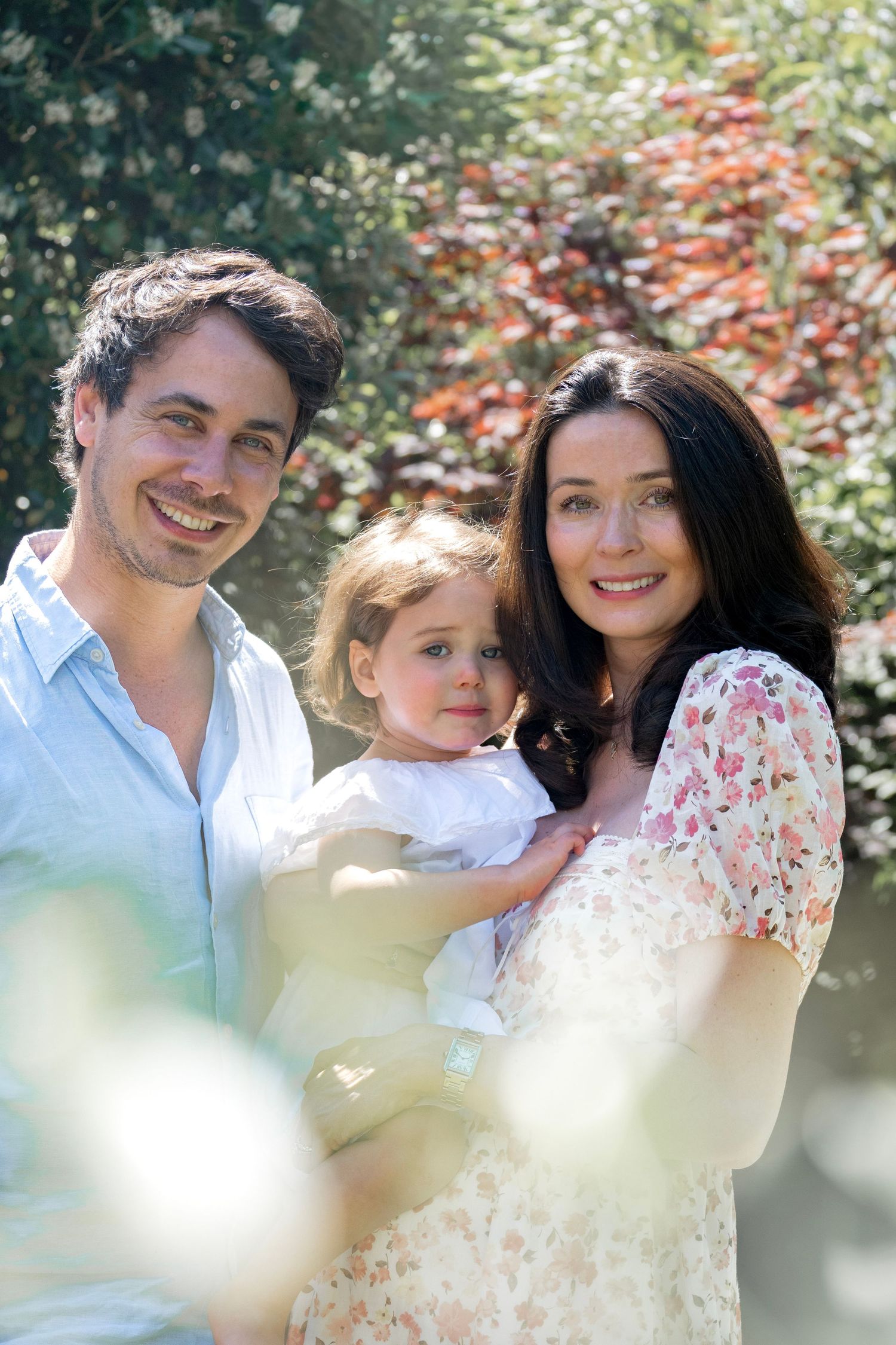 Happy family portrait of parents holding toddler daughter outdoors in sunny garden with colorful foliage.