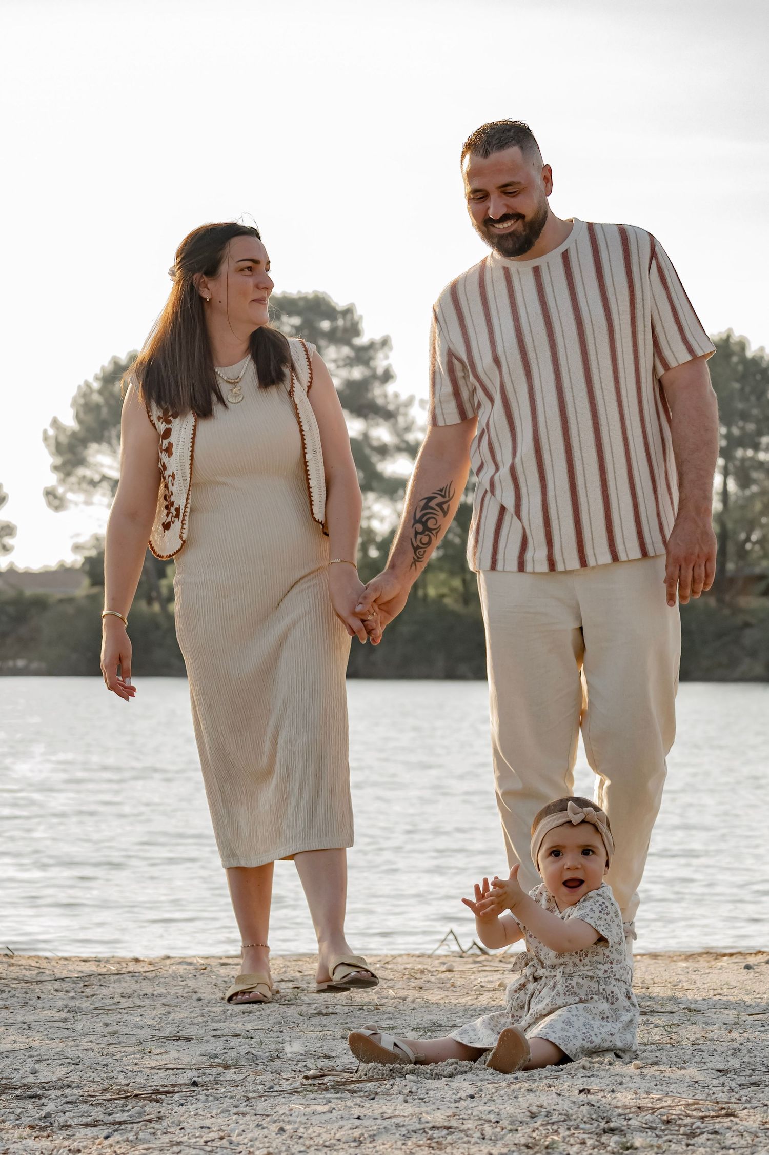A happy family with a baby sitting on a sandy beach by a lake, parents holding hands in neutral tones.