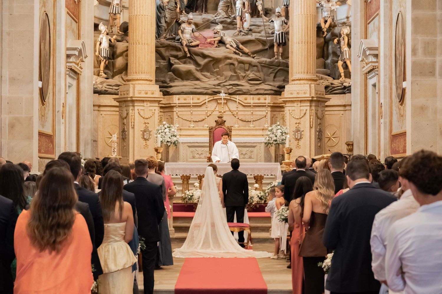 Elegant church wedding ceremony with bride and groom at ornate altar, guests seated in pews, red carpet aisle.