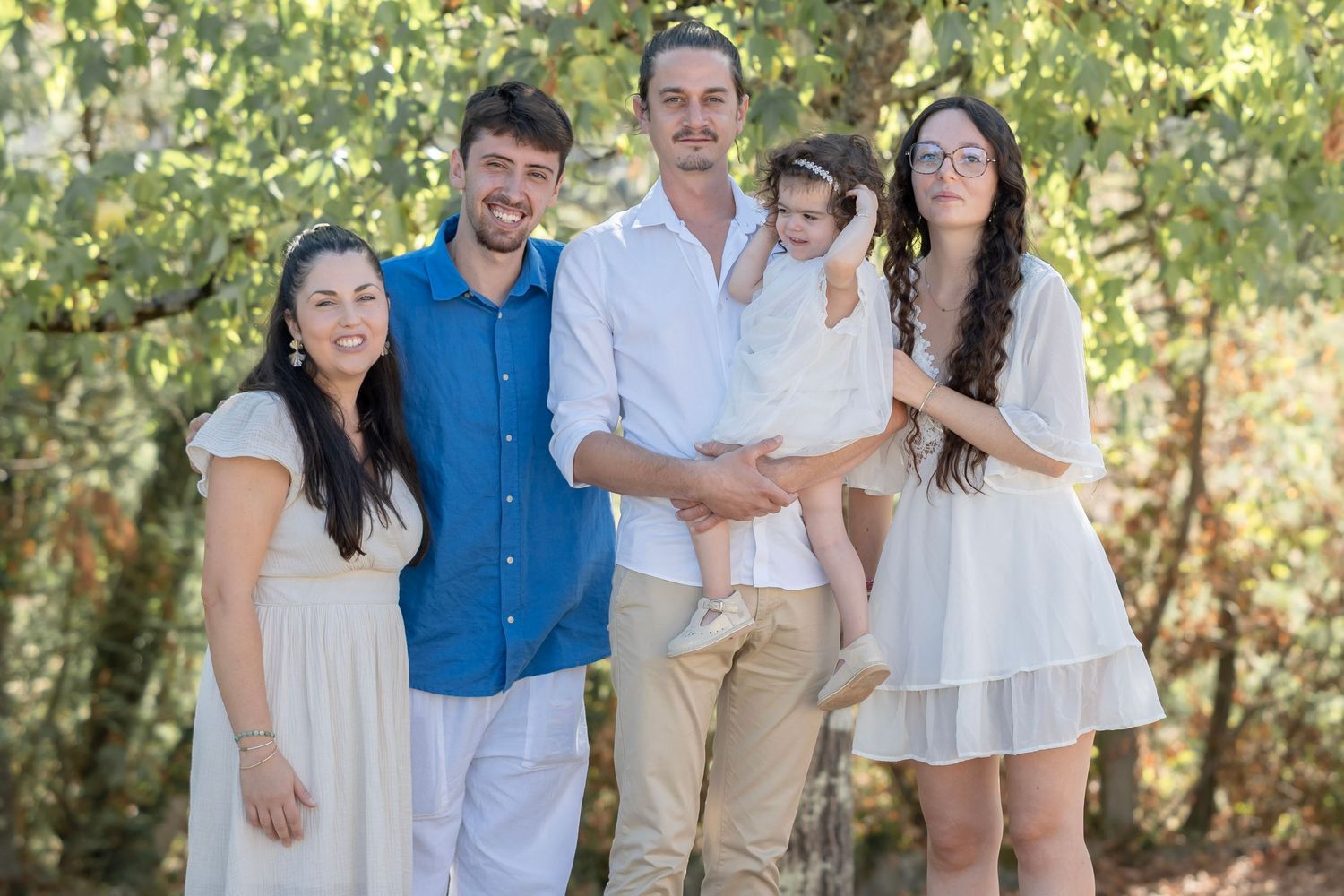 A group of five people wearing coordinated white and blue outfits pose together outdoors in front of greenery.