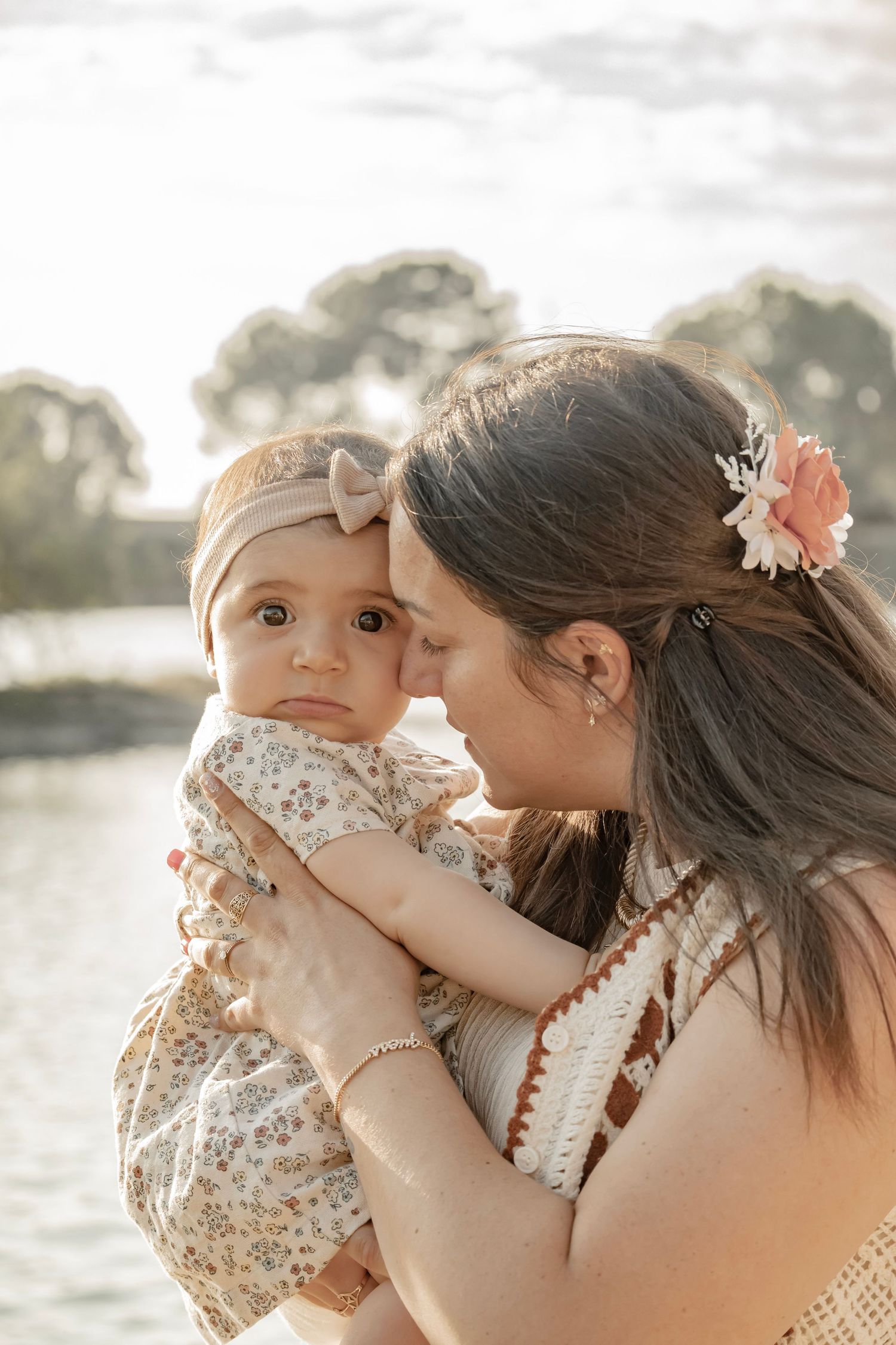 A mother tenderly kisses her baby girl outdoors, both wearing floral accessories near a sunlit lake.