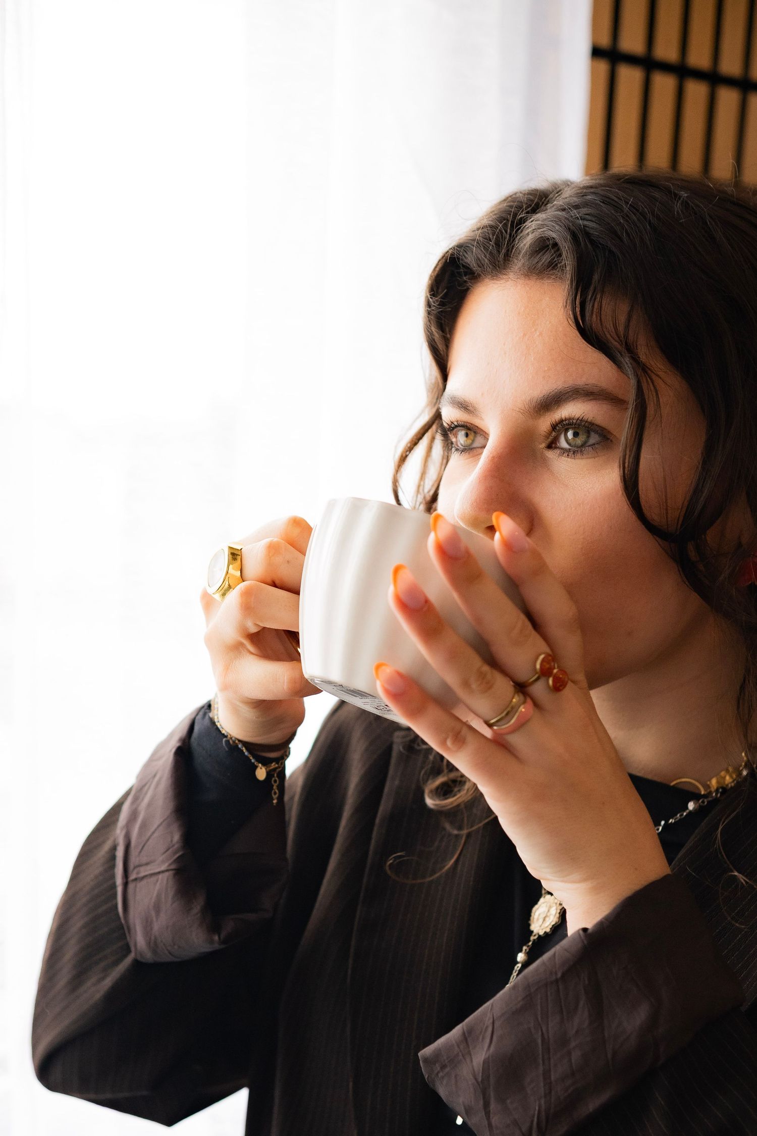 Woman with dark hair sipping from a white espresso cup, wearing gold rings and a dark blazer near a bright window.