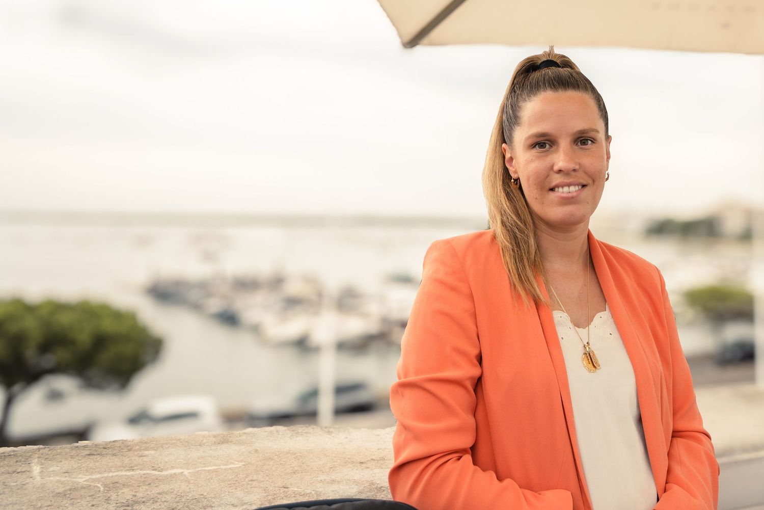 Professional wearing a coral blazer and cream top poses outdoors against a blurred natural background.