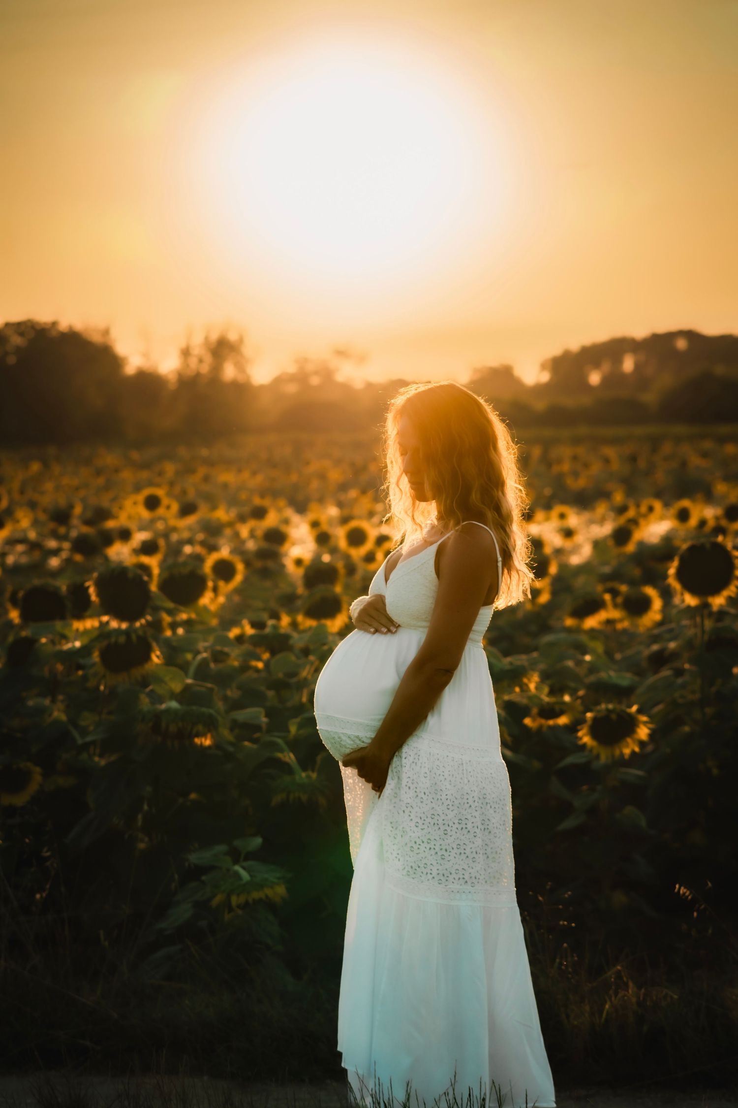 Pregnant woman in white dress standing in sunflower field at golden sunset, cradling her baby bump.