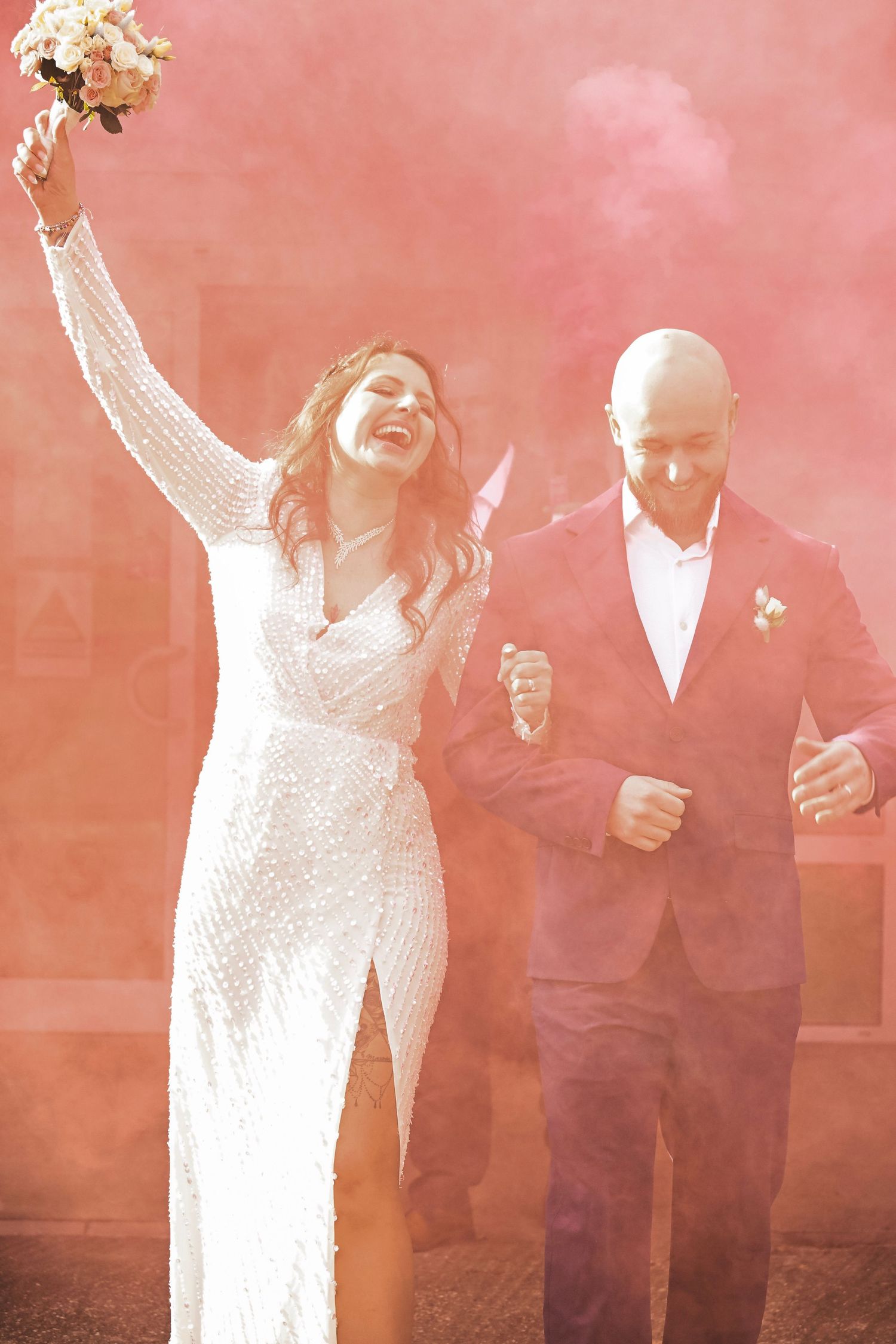 Newlyweds celebrate through pink smoke with raised bouquet and formal attire.
