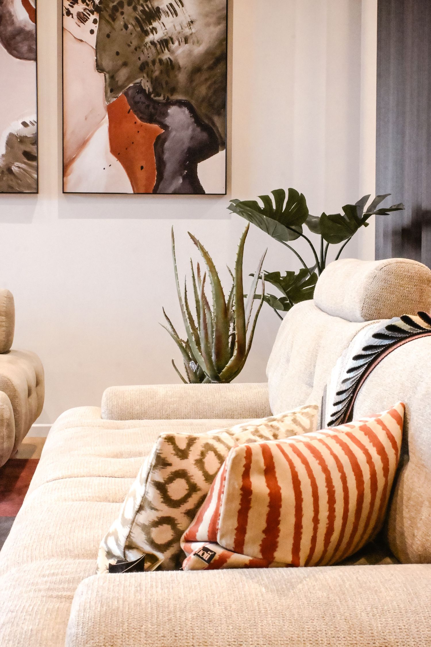 Modern living room corner with beige couch, decorative orange striped pillows, and monstera plant near abstract wall art.