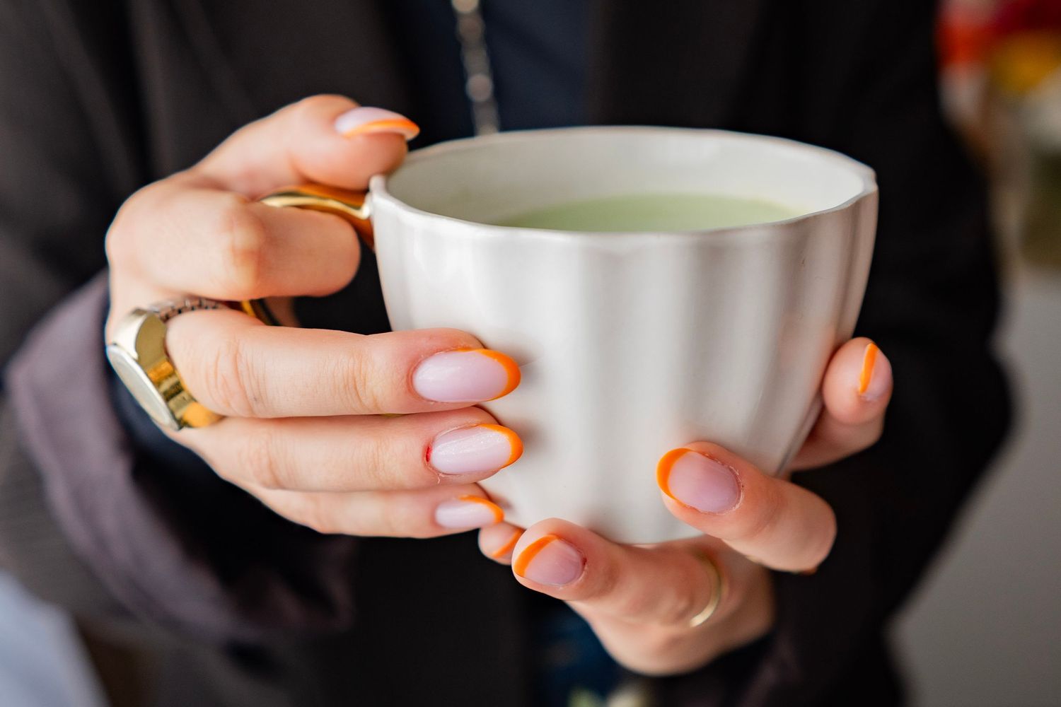 Close-up of hands with orange-tipped nails holding a white ceramic cup of green matcha tea.