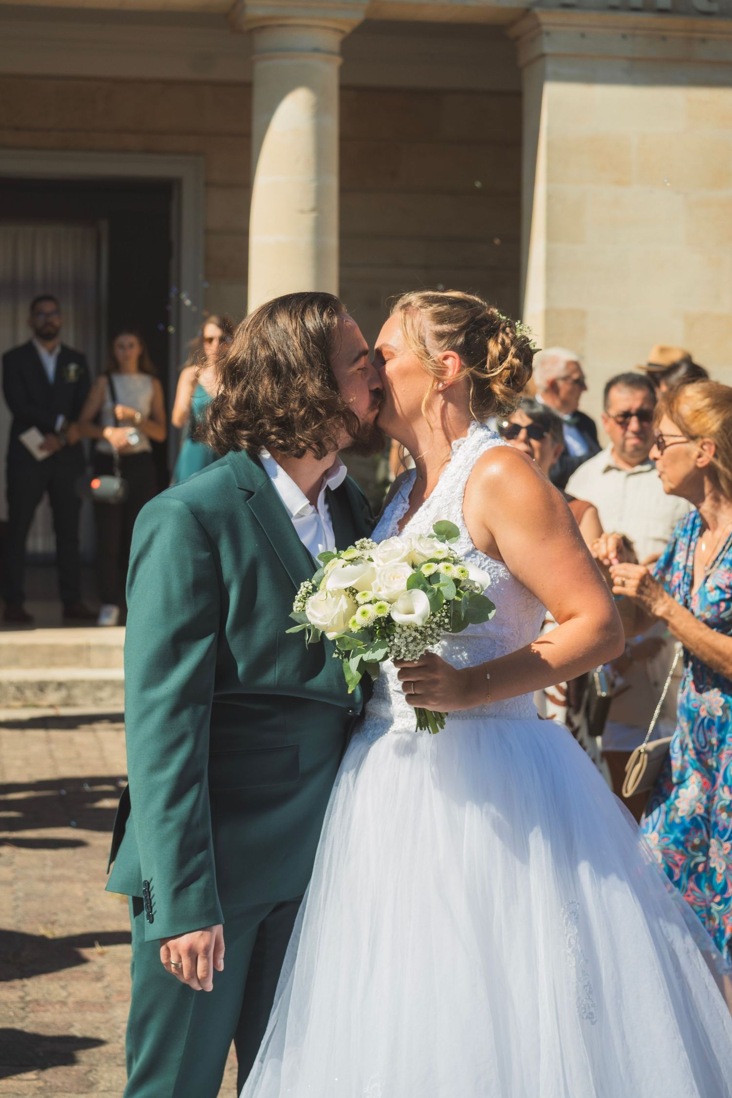 Bride and groom share a kiss outside a venue, bride holding white bouquet, guests watching in background.