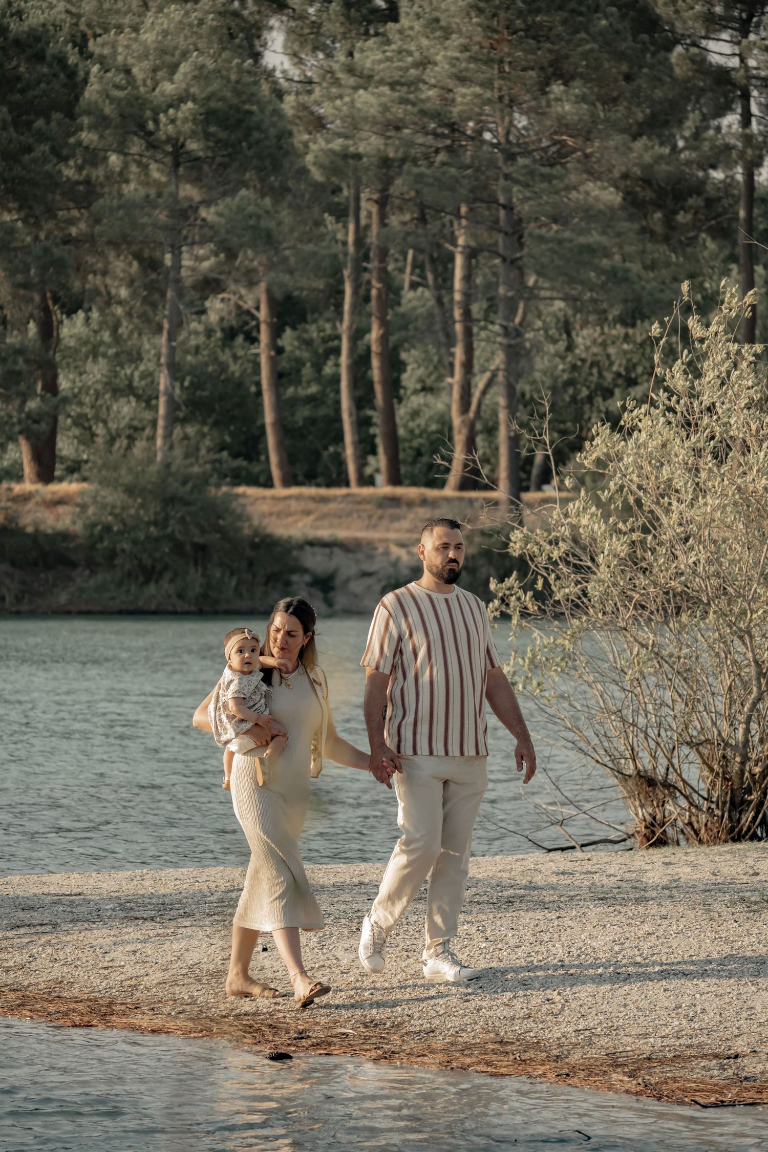 Young family with baby strolling along a scenic lakeside path surrounded by pine trees in warm natural light.