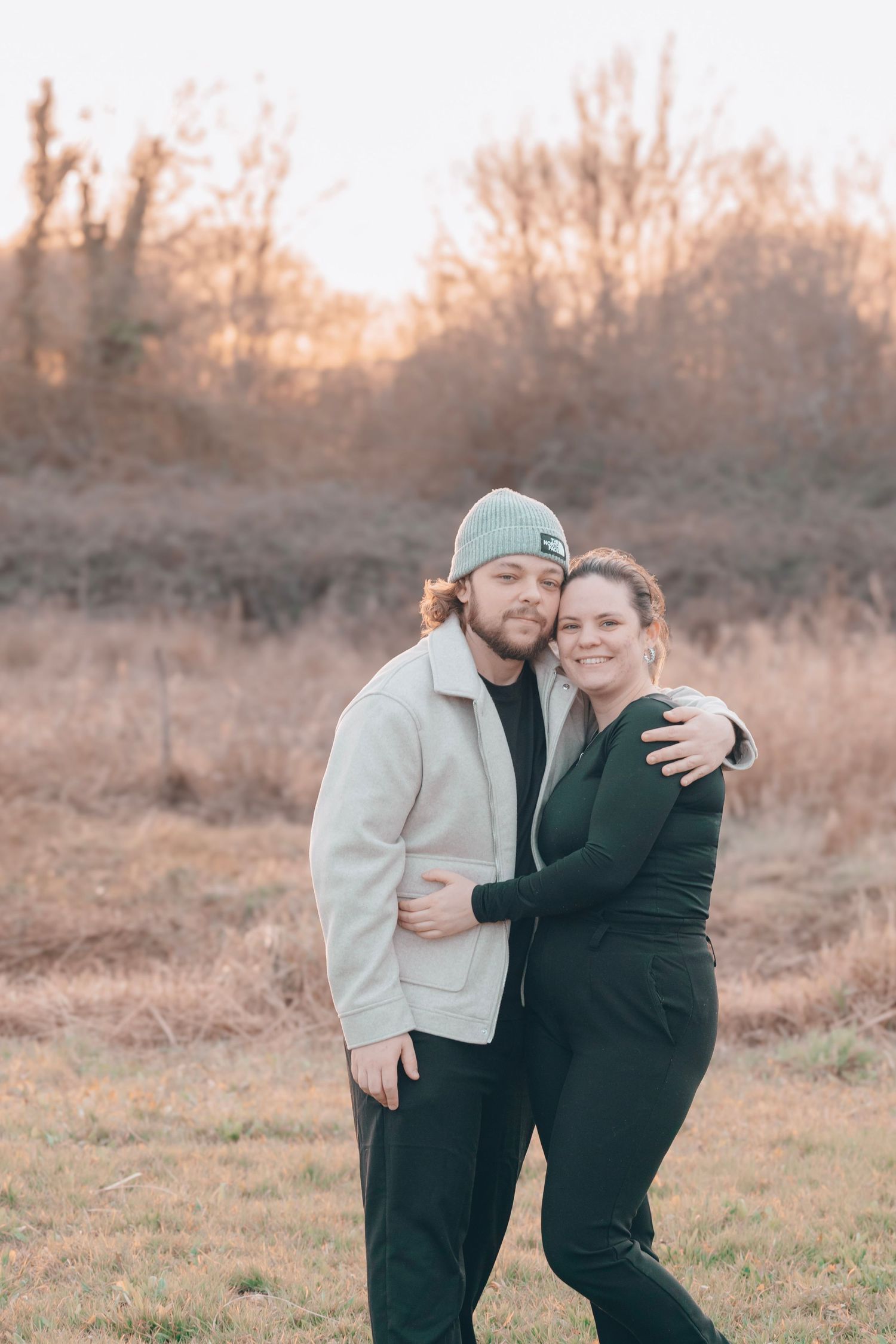 Couple in casual winter wear embrace in a golden sunset field.