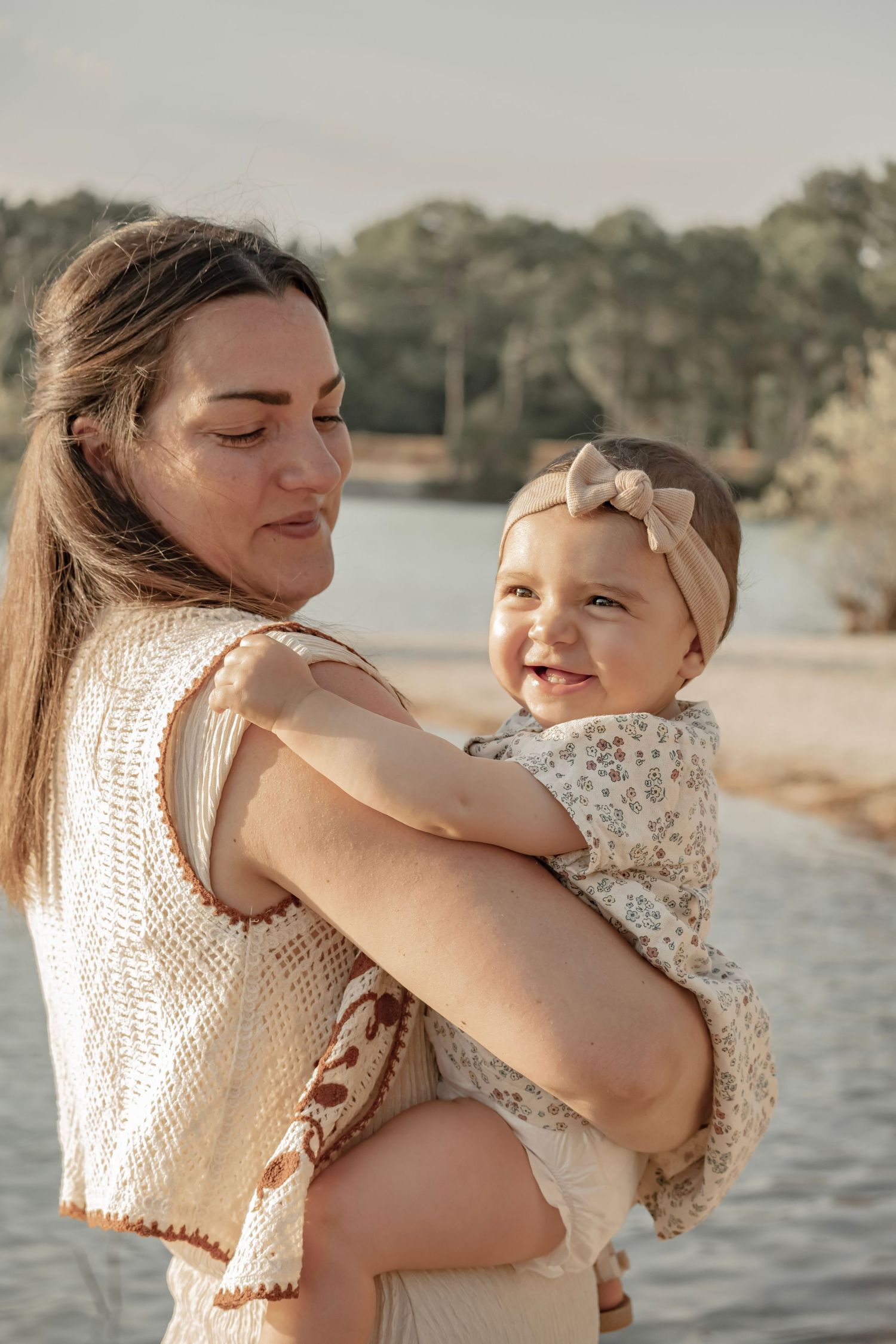 A smiling baby in a floral outfit and bow headband being held by a young woman near a sunlit lake.