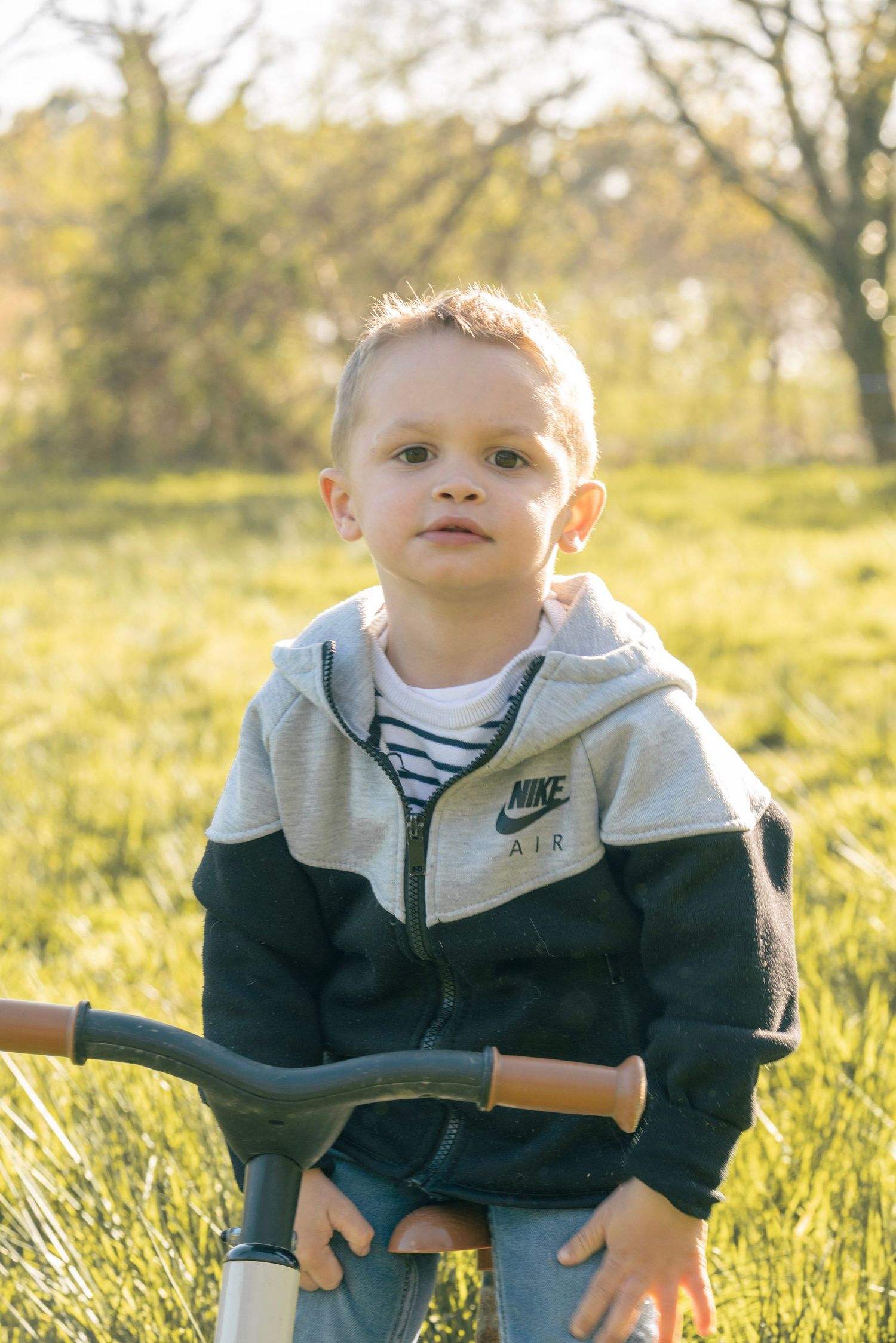 Young child wearing a grey and navy Nike Air hoodie stands in sunlit grass field during golden hour.