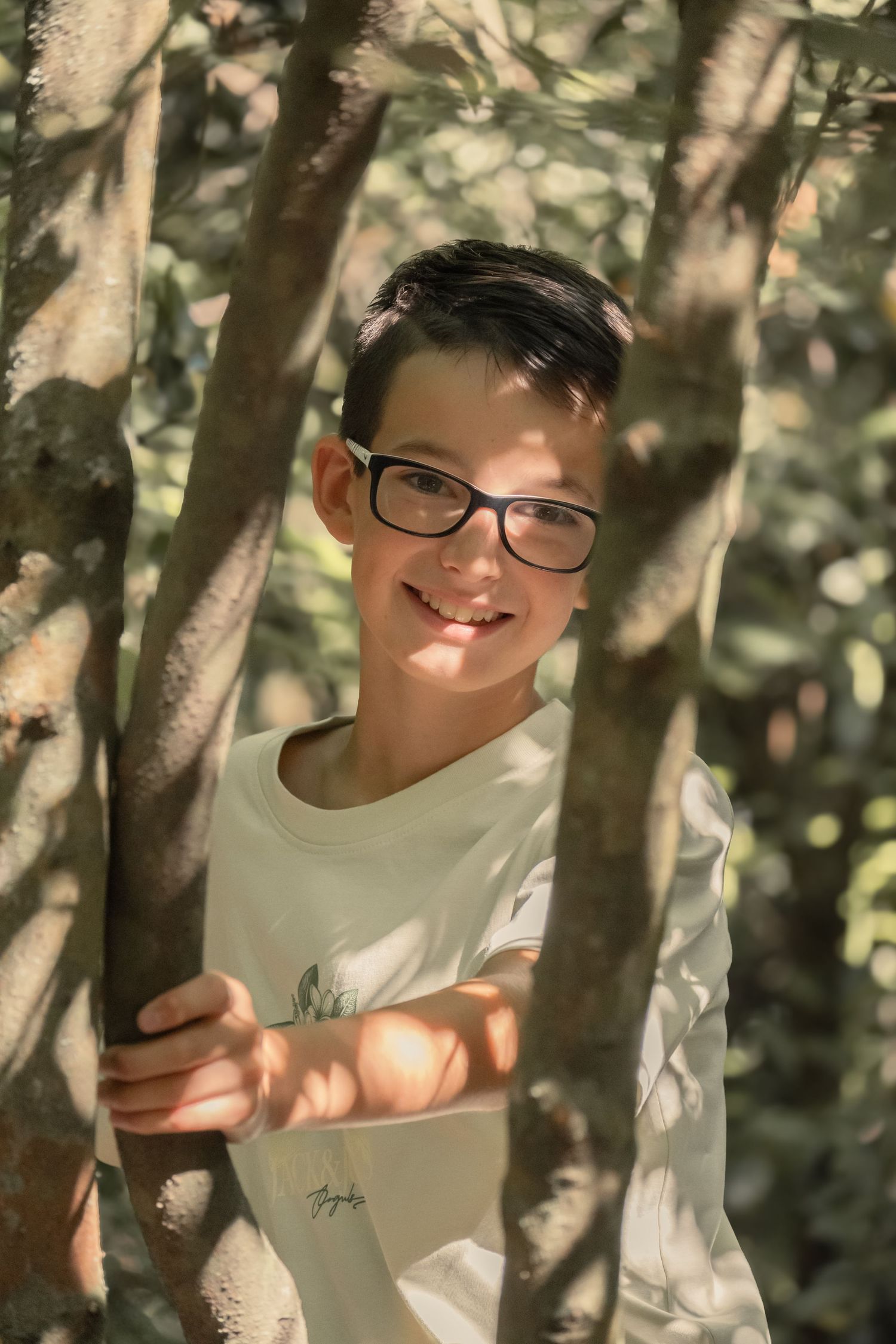Young person wearing glasses and white shirt stands amid tree branches in dappled sunlight.