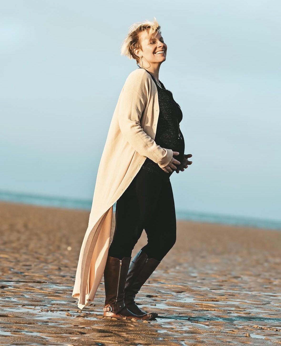 Person standing on a beach at sunset wearing a long beige cardigan over black outfit, looking away from camera.