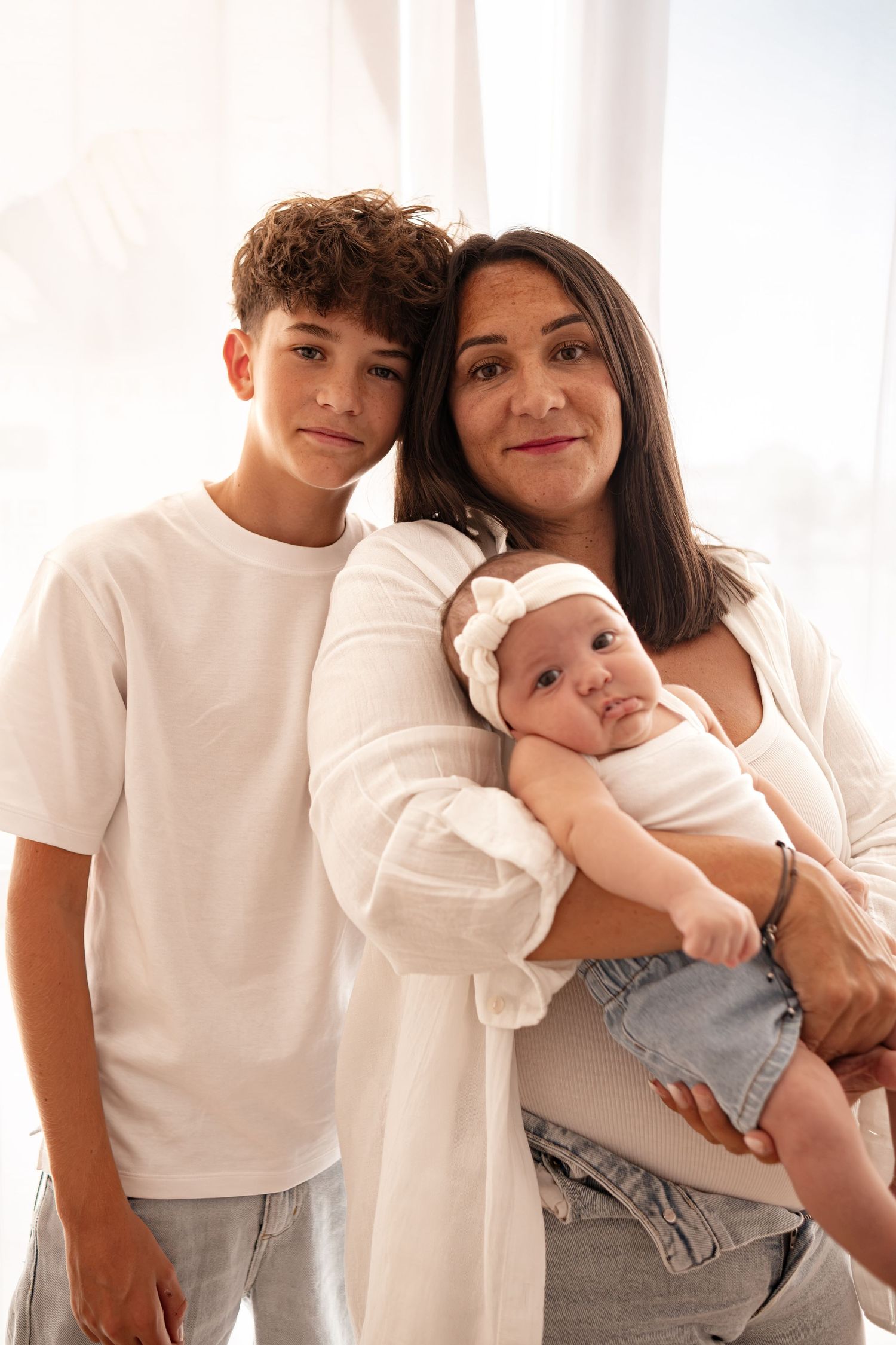A mother holds her baby girl while her teenage son leans in close for a family portrait in white outfits.