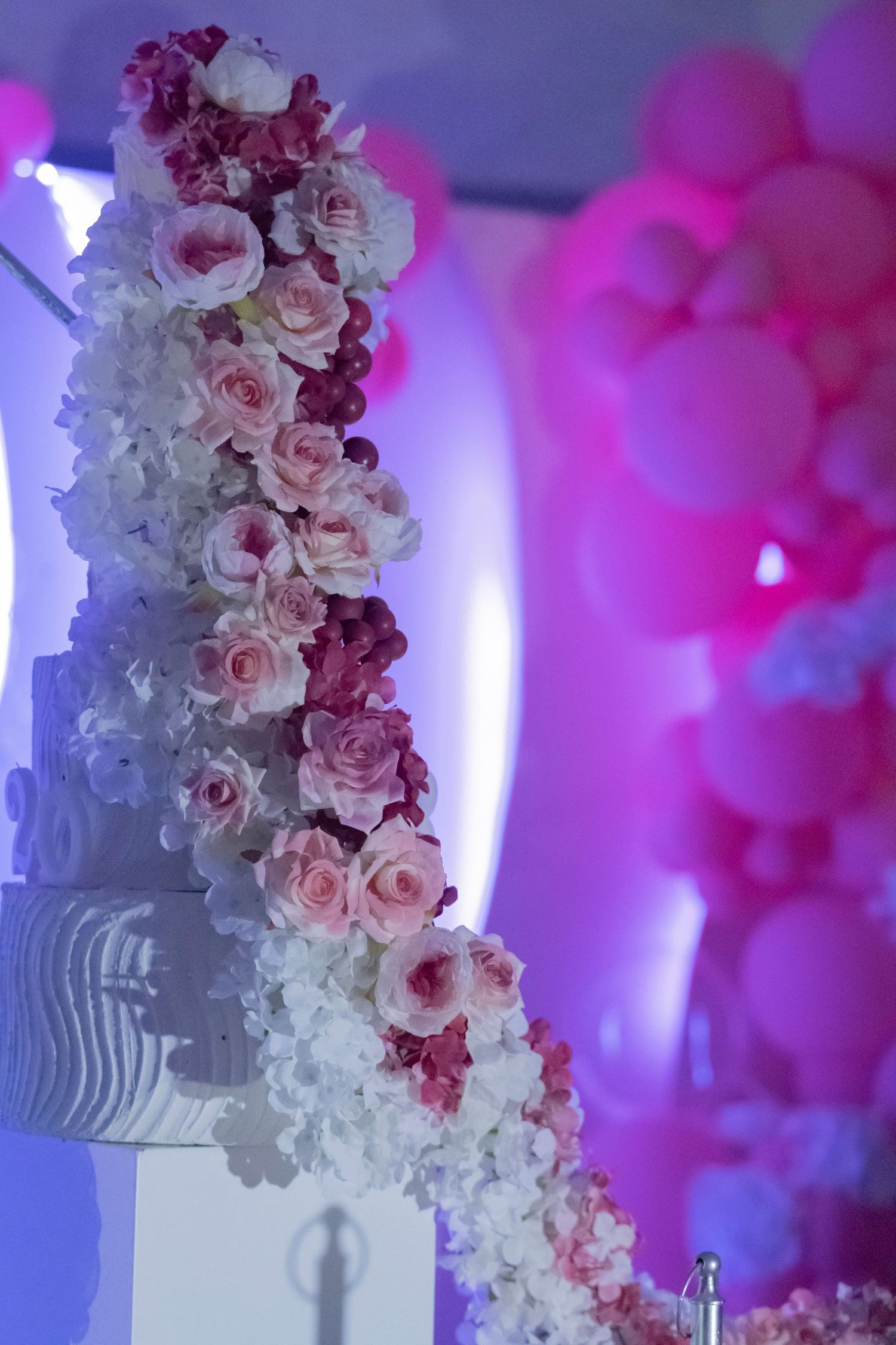Elegant white wedding cake decorated with cascading pink roses and orchids under pink and purple ambient lighting.