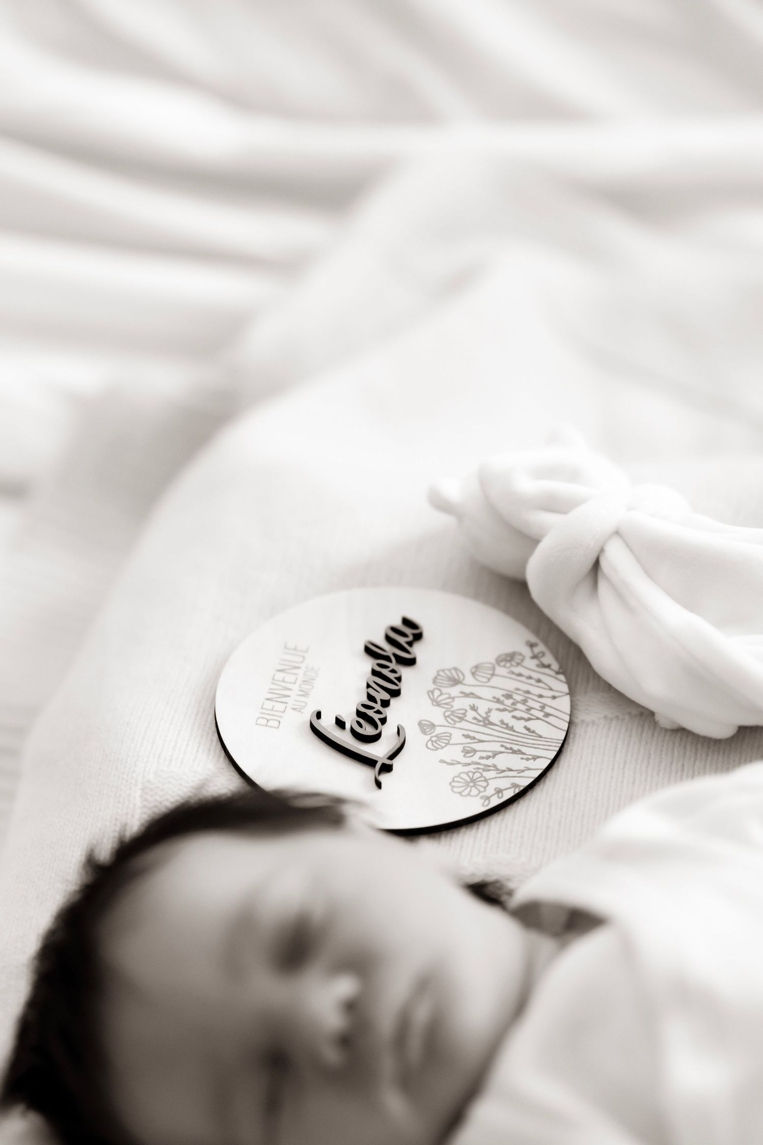 Black and white newborn photo with a wooden name disc reading Liambu beside a sleeping baby wrapped in white cloth.