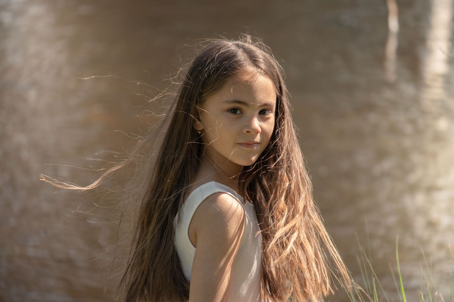 A young person with long brown hair wearing a white sleeveless top poses against an outdoor blurred background.