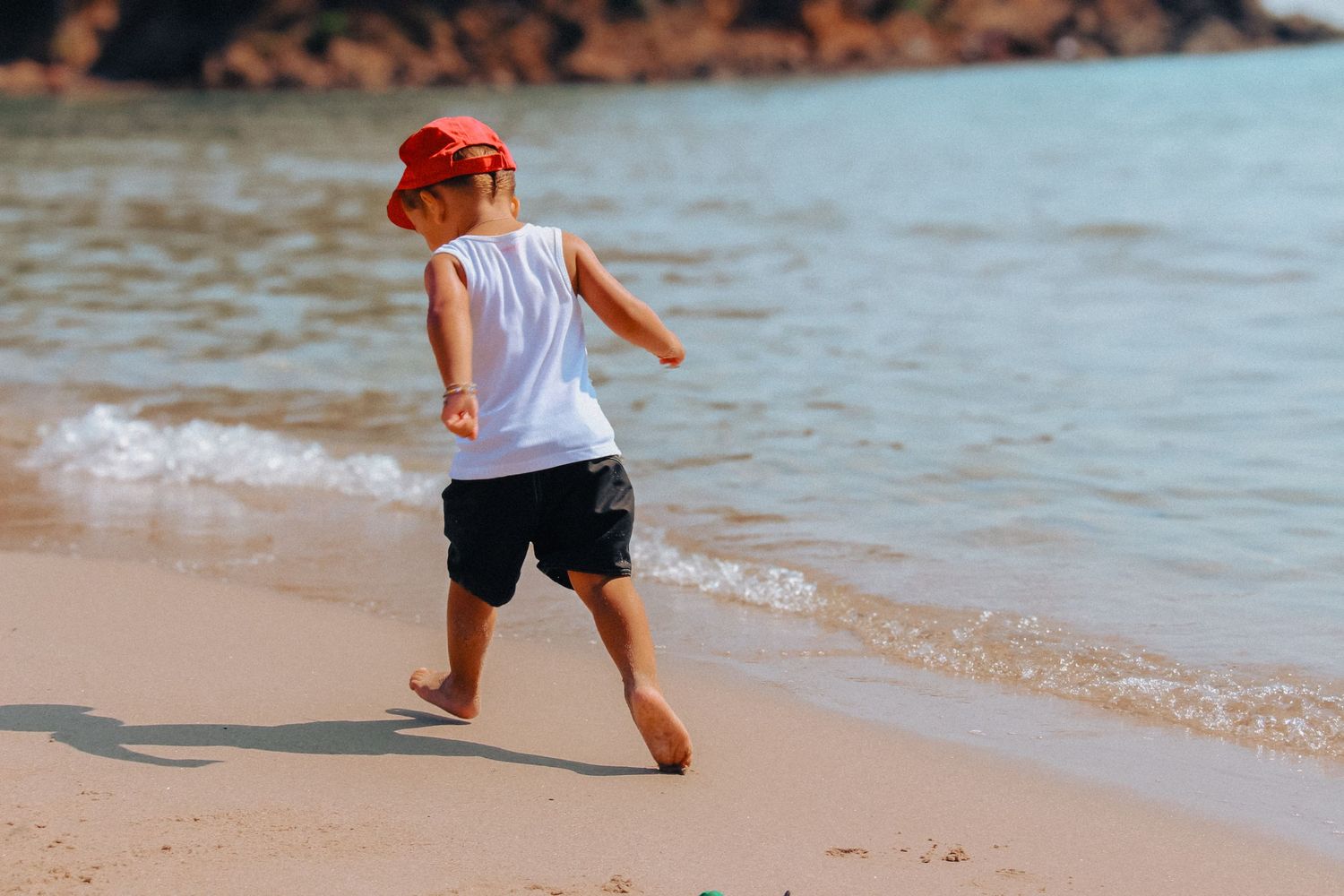Small child wearing a red hat runs along the sandy beach shore with water in the background.