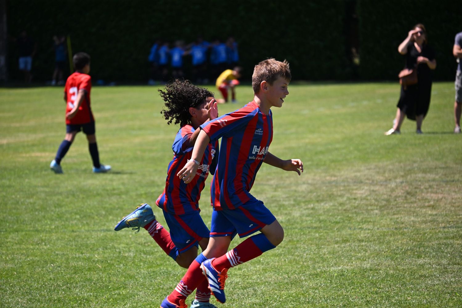 Two youth soccer players in red and blue striped jerseys run side by side during an outdoor match on a grass field.
