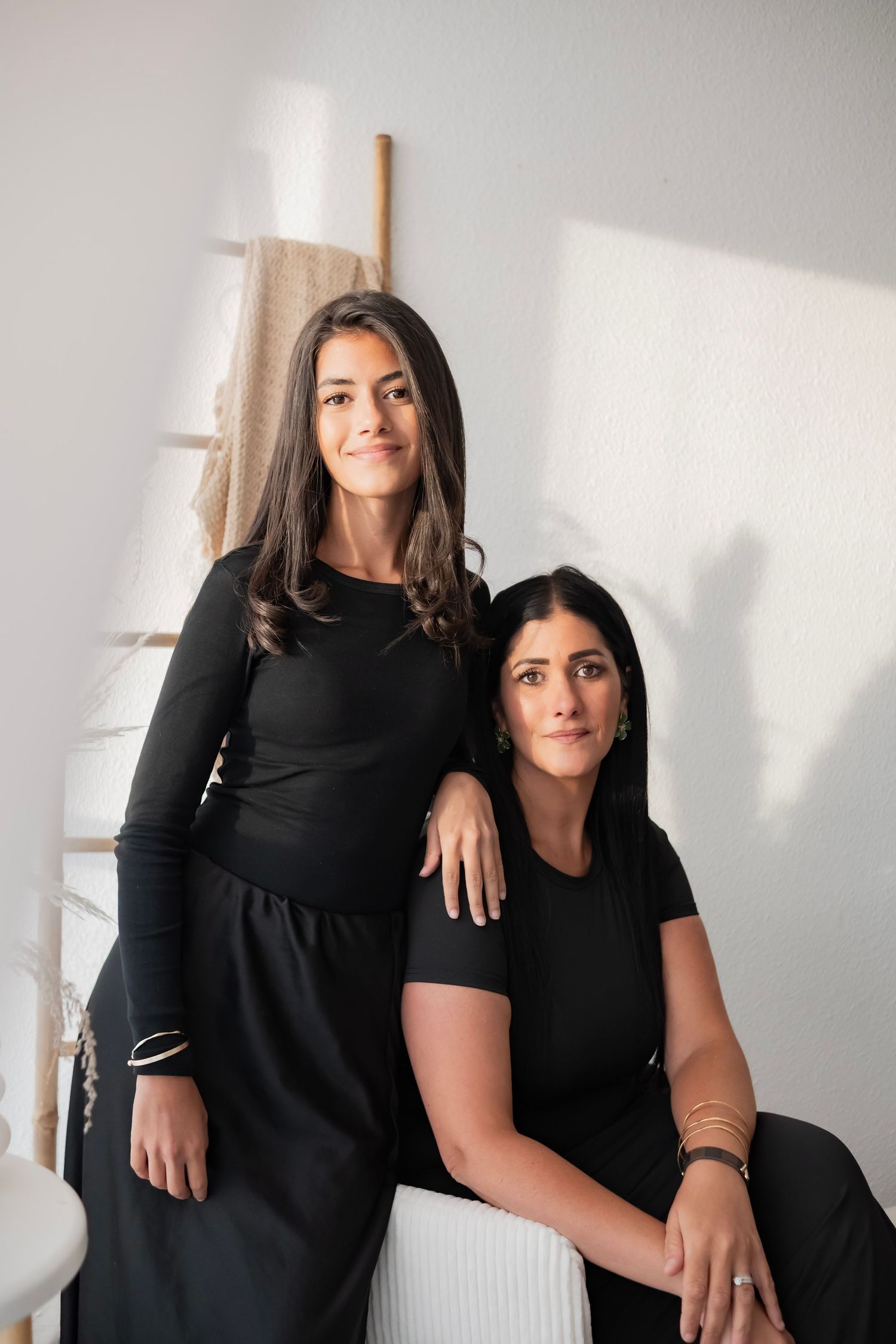 Two women in matching black outfits pose together in a bright, minimalist studio setting.