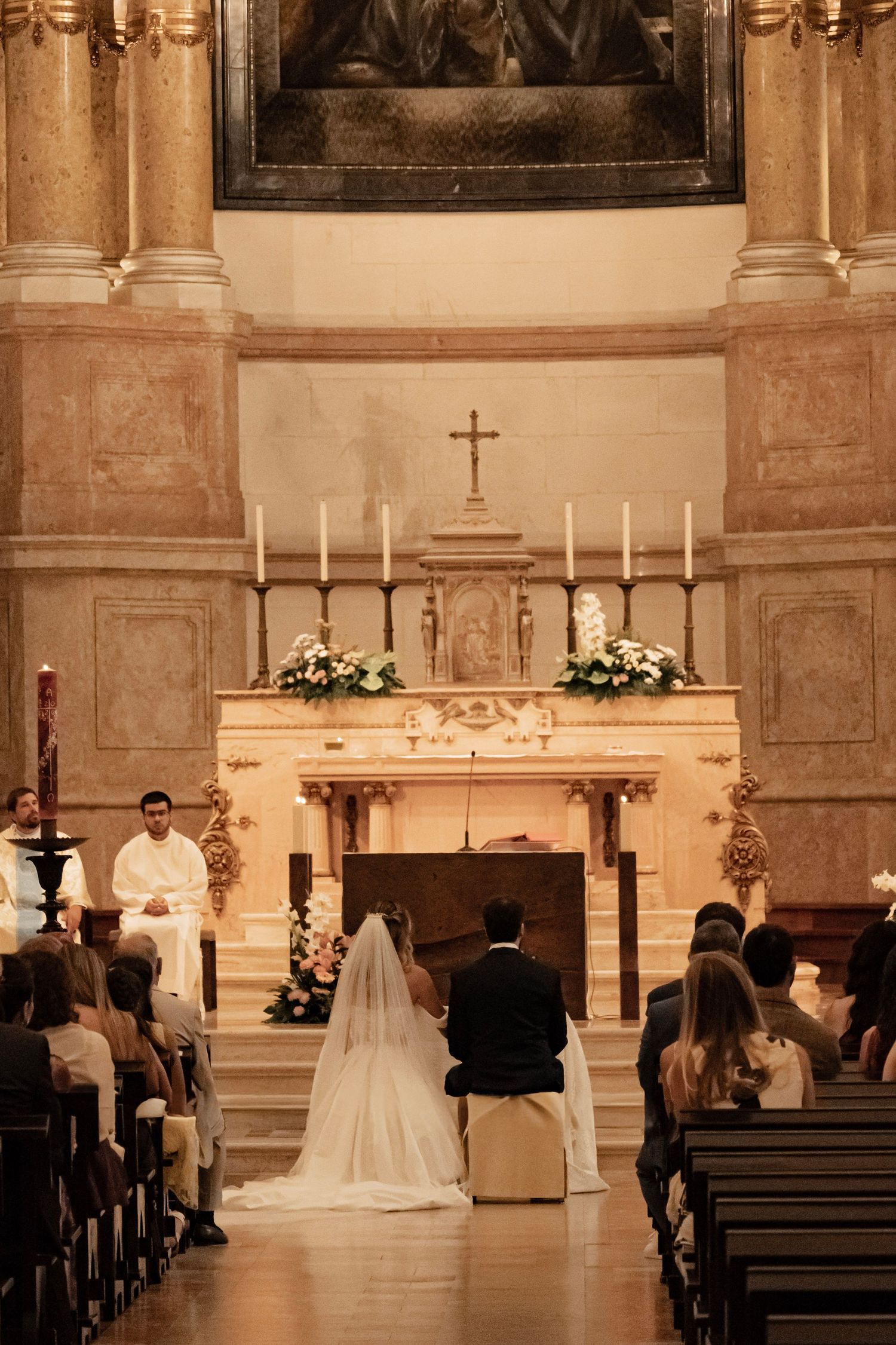 Bride and groom kneeling at church altar during wedding ceremony with candles and cross in ornate chapel.