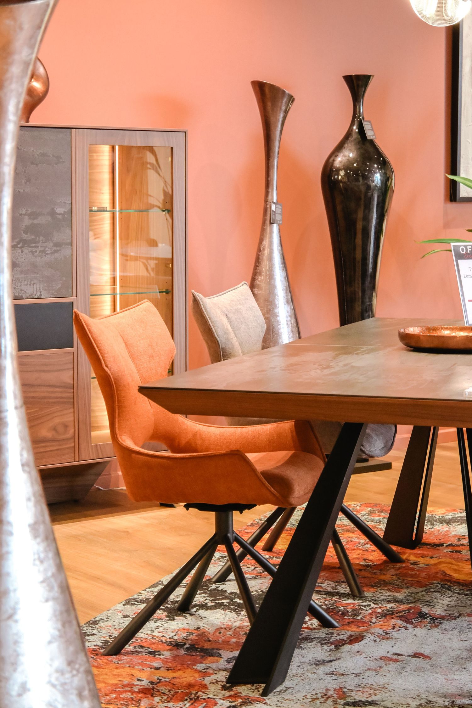 Modern dining area with orange leather chair, wooden table, metal legs, and tall decorative vases against coral walls.
