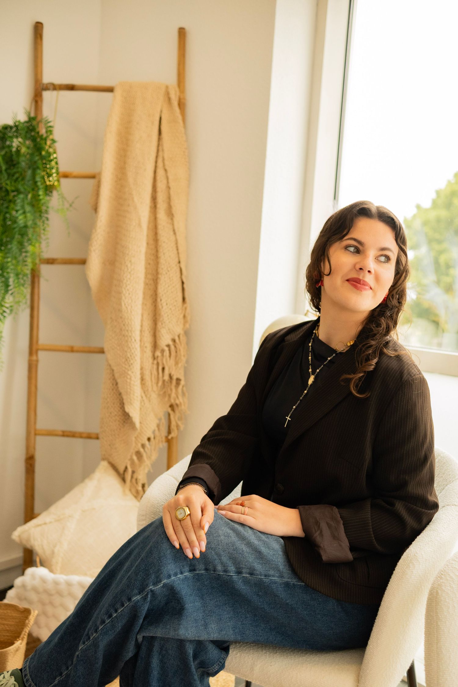 Woman in black blazer and jeans sits in white chair near window, with decorative ladder and blanket in background.
