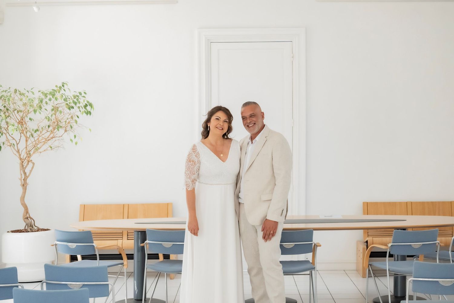 Bride and groom posing together in a bright white venue with blue chairs and a potted tree.