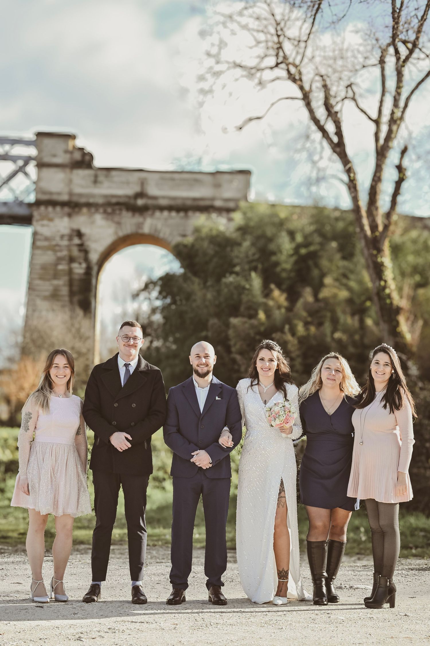 Wedding party poses in formal attire in front of a stone arch bridge.
