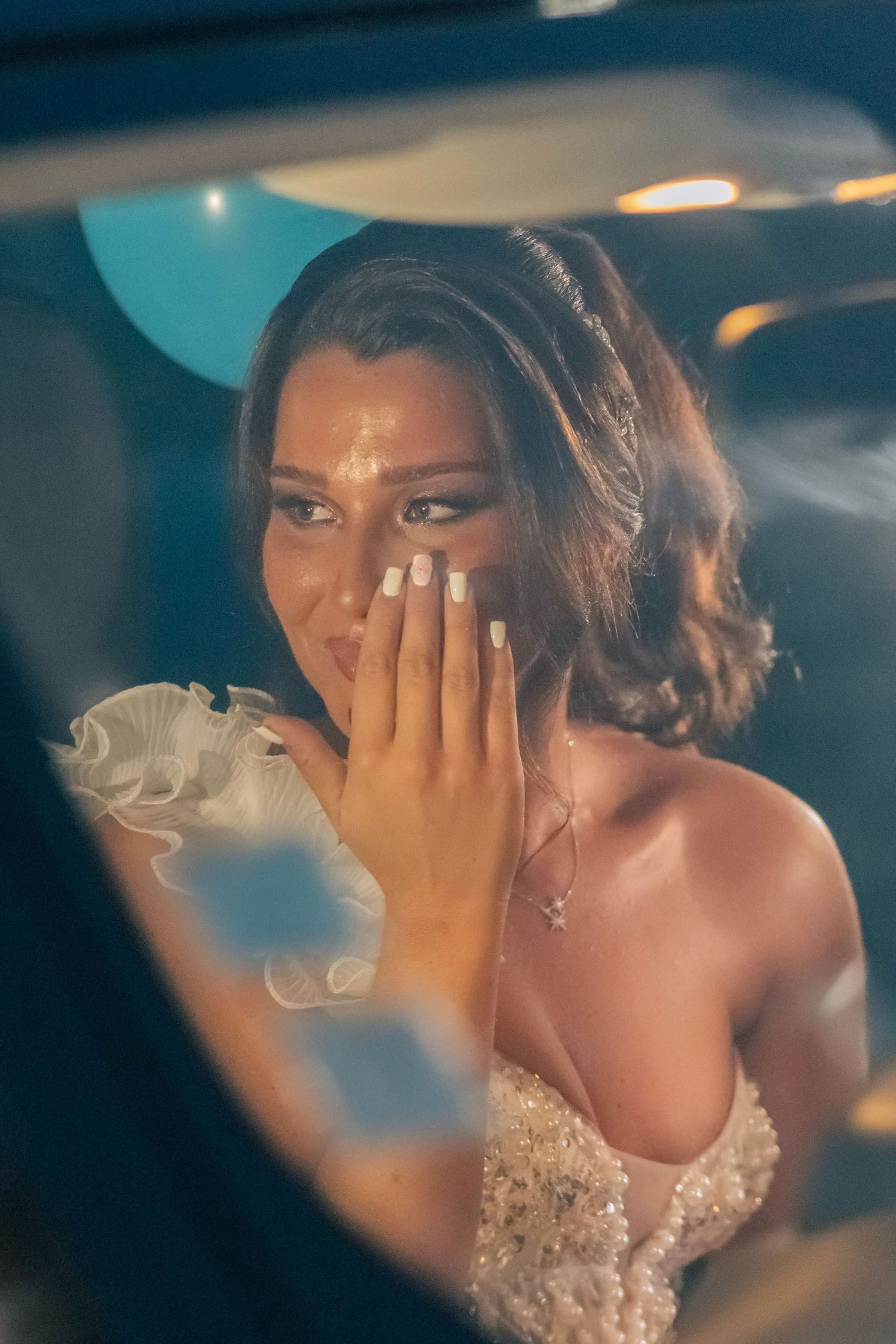 In the car's reflection, someone wearing an elegant white lace bridal gown dabs tears from their eyes in an emotional moment.