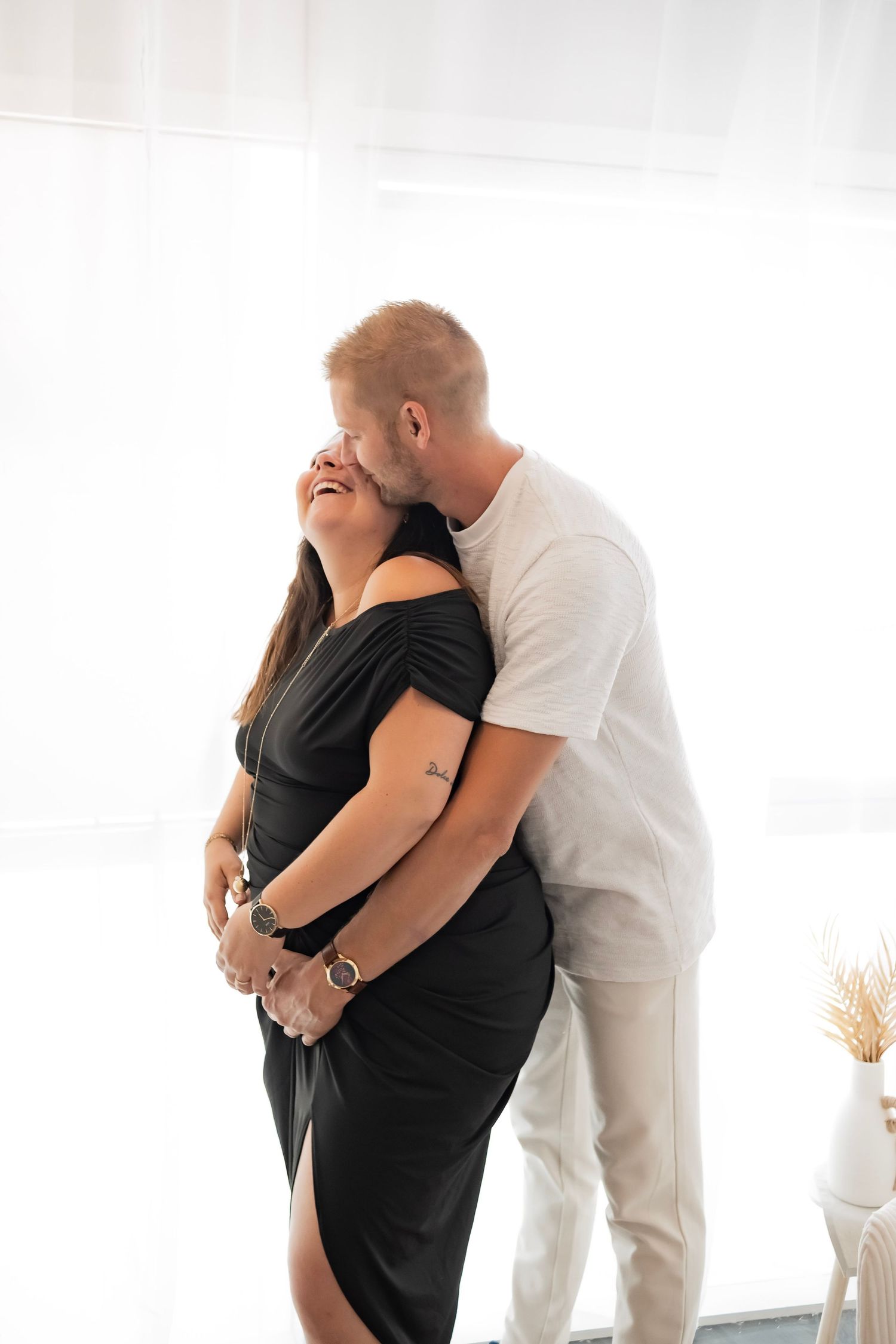 A man in white kisses a smiling woman in a black dress while embracing her from behind indoors.