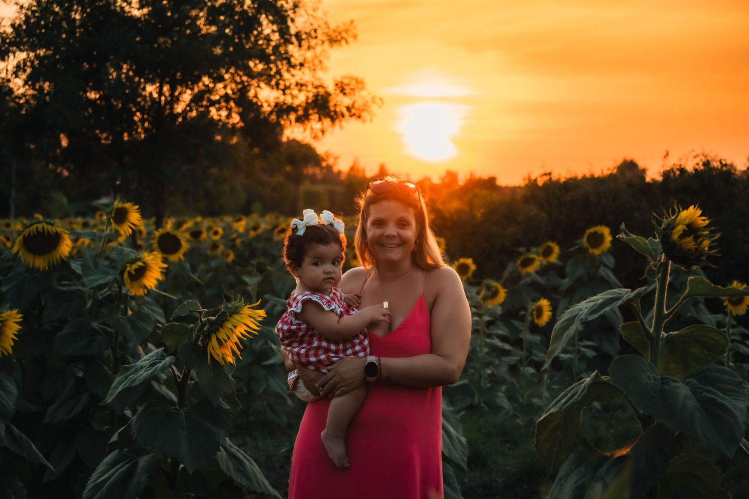 Woman in pink dress holds baby girl in sunflower field at golden sunset.