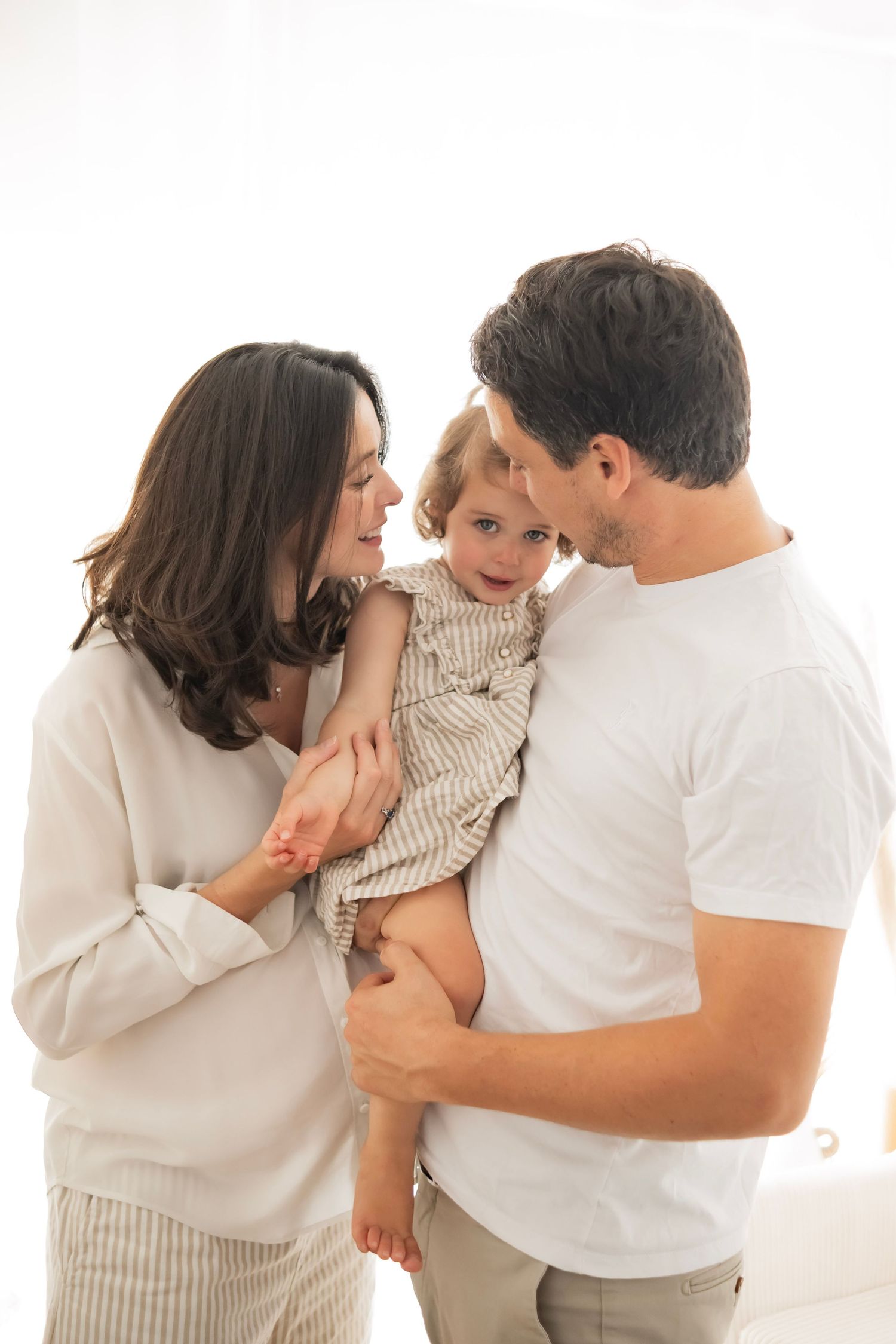 Happy young family with toddler daughter, parents smiling at child in bright white studio setting.
