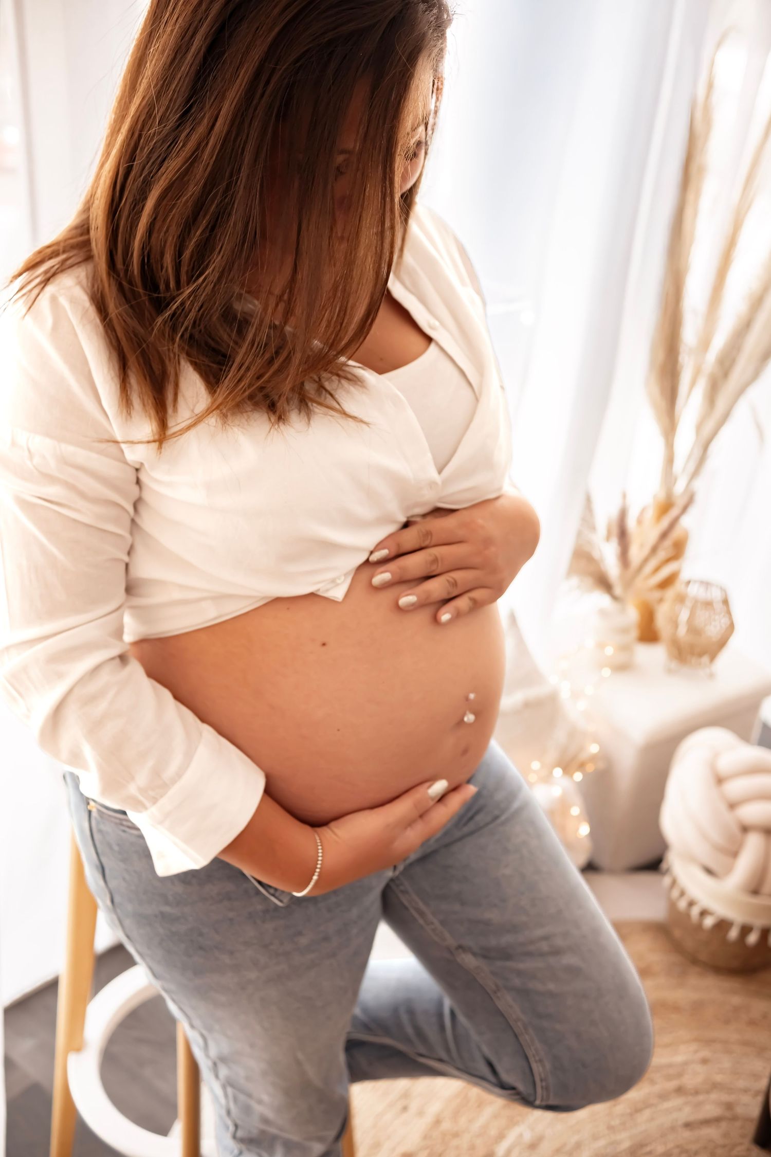 Pregnant woman in white shirt and jeans cradling her baby bump while sitting, with soft natural light in background.