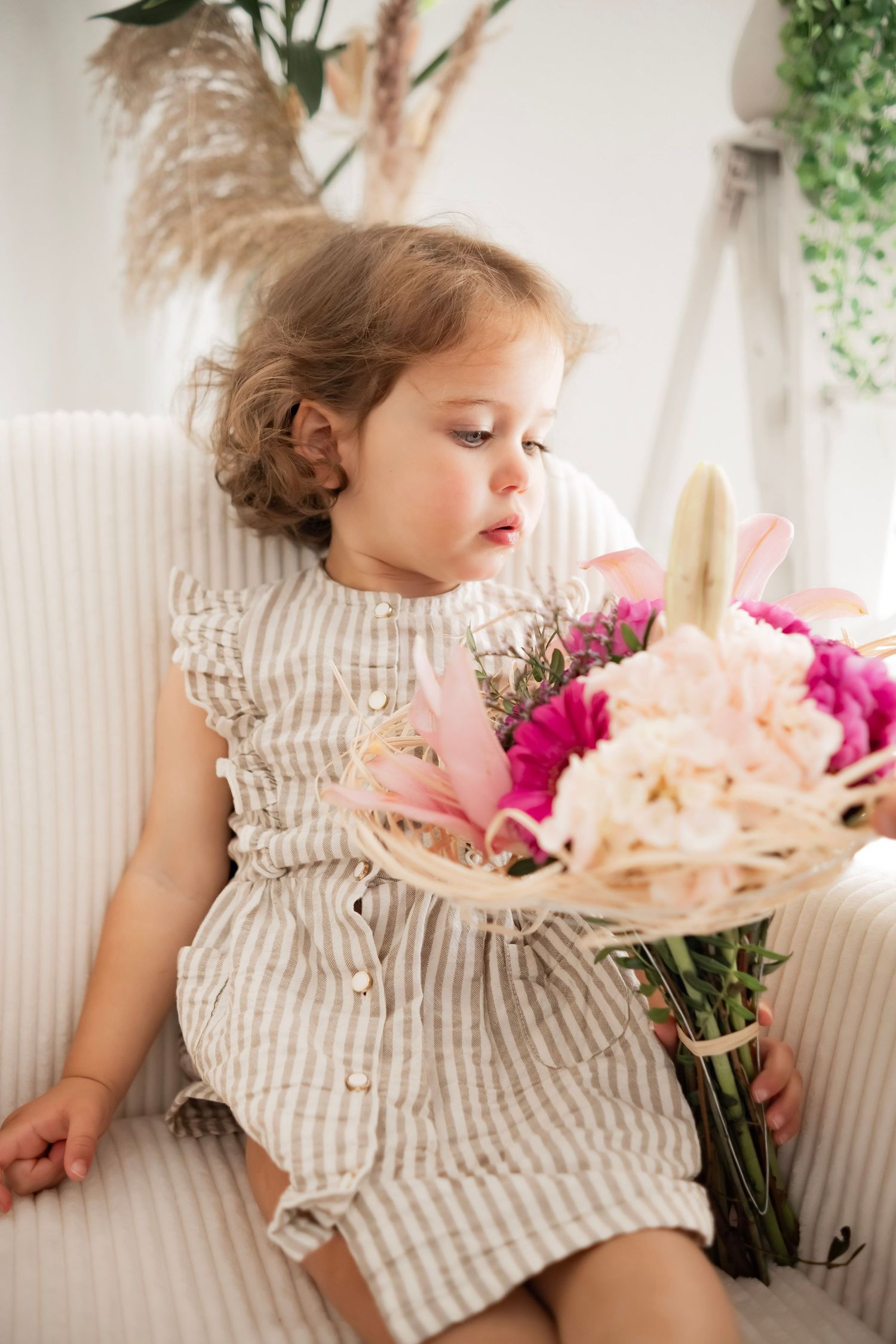 Toddler girl in striped dress holding a bouquet of pink and white flowers while sitting in a white chair.