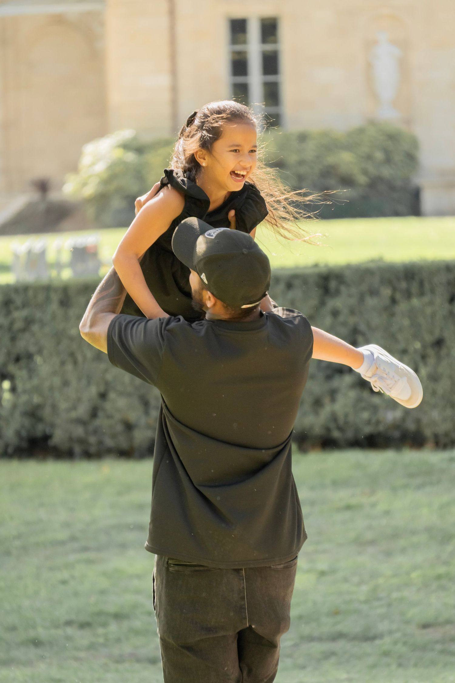 A couple embraces joyfully in an outdoor setting with green grass and hedges in the background.